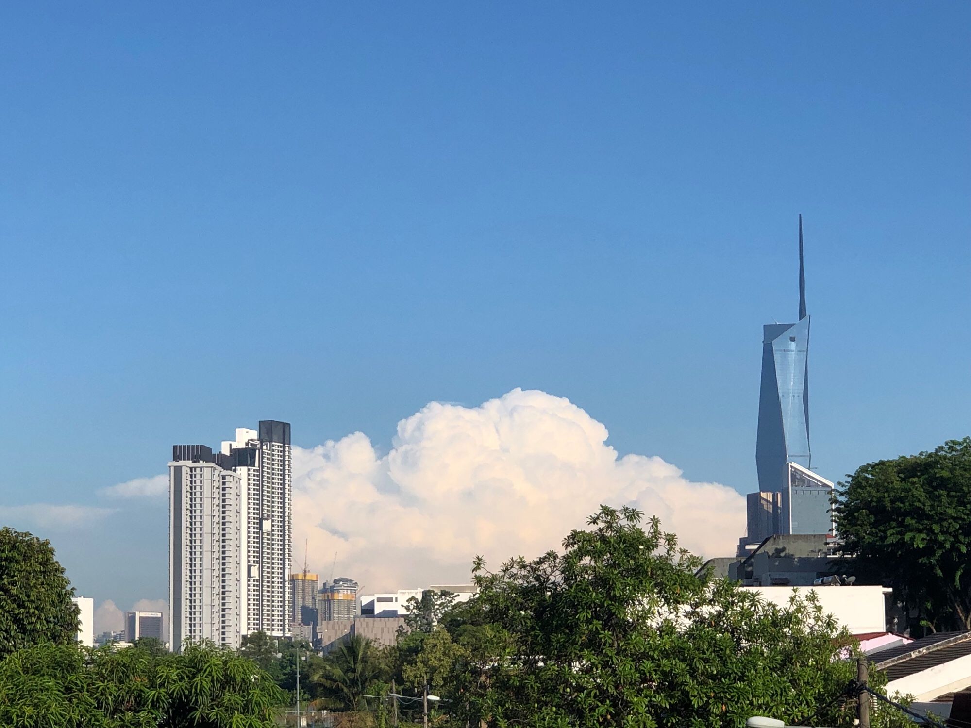 Blue sky with some clouds, Warisan Merdeka Tower and trees in the foreground. 