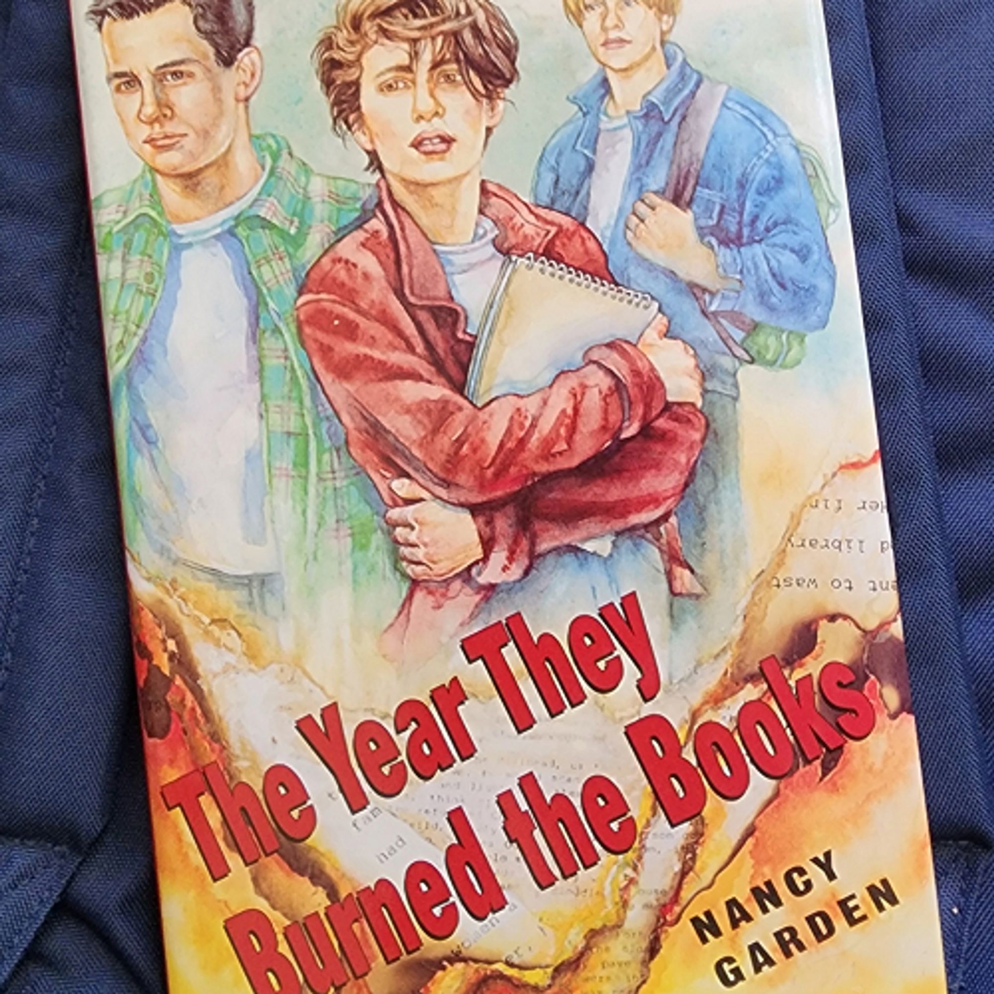 The Year They Burned the Books by Nancy Garden cover an illustration by Alexa Rutherford of three high school students one clutching a notebook, the book title in red over burning pages with text.