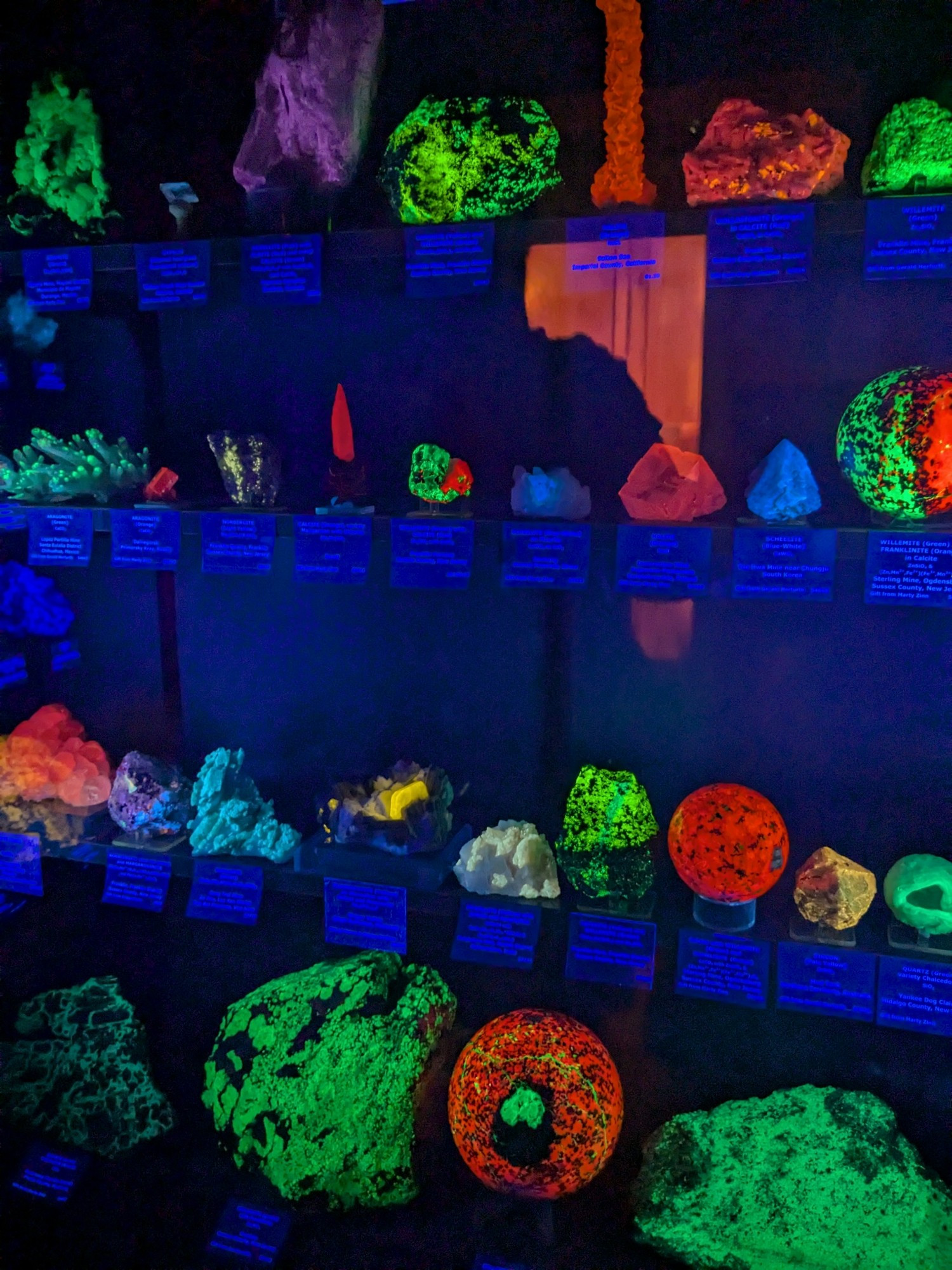 a wall of various fluorescent crystals under a blacklight making them shine in orange, green, blue, and purple hues