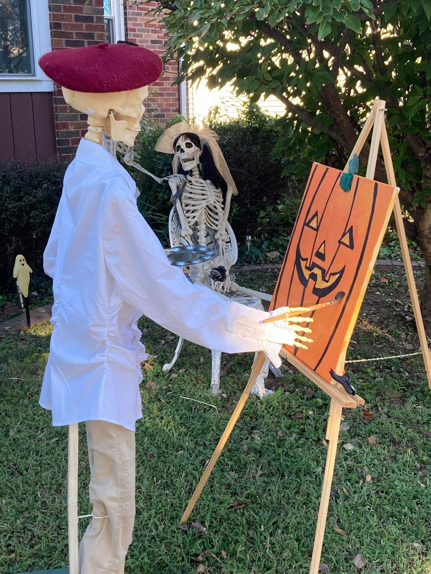 A plastic skeleton dressed as an artist. It is wearing a beret and holding a paintbrush which it is using to paint a Jack-o-lantern on the canvas in front of it.