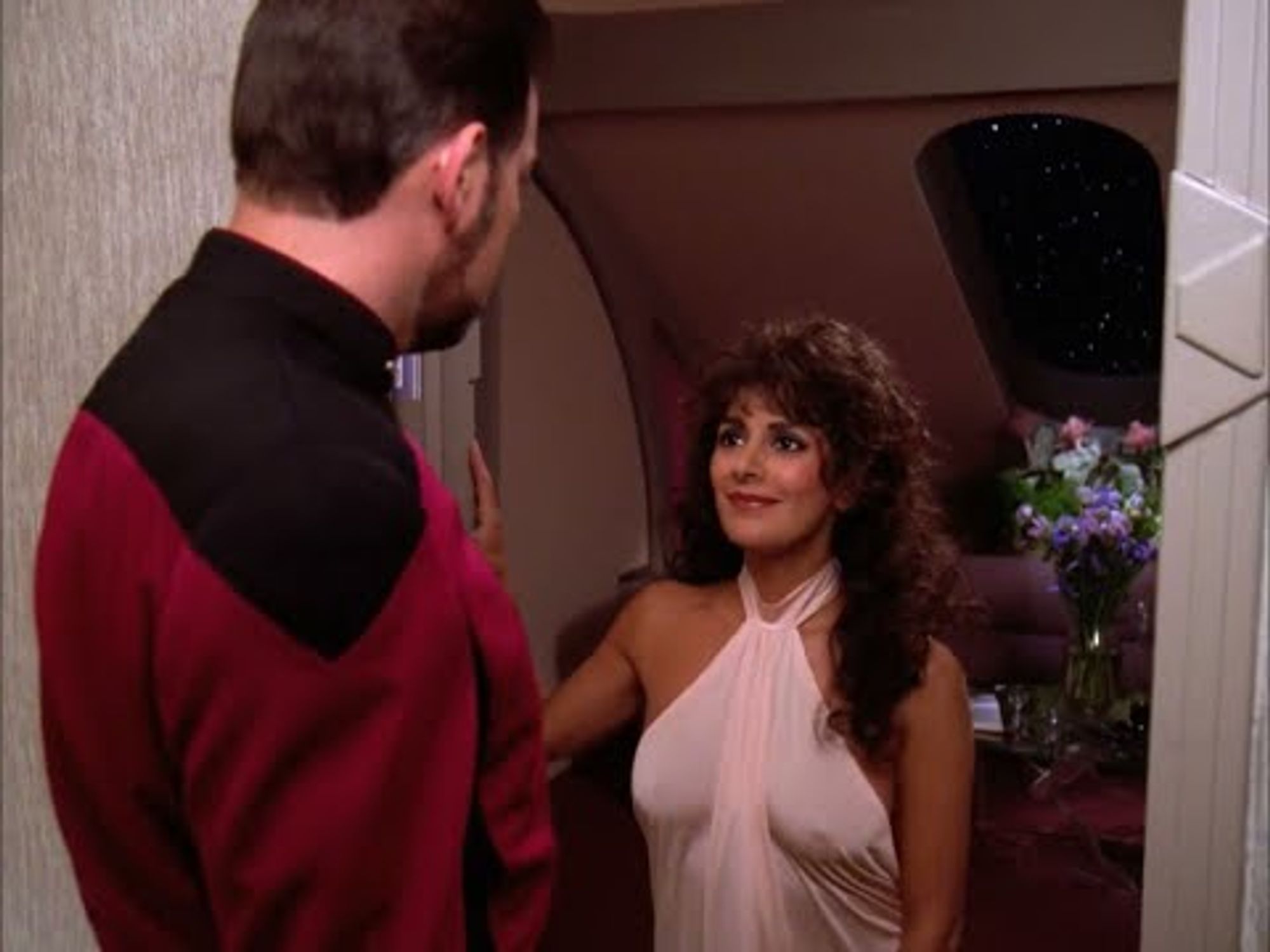 Troi greets Riker at her door wearing a white dress.