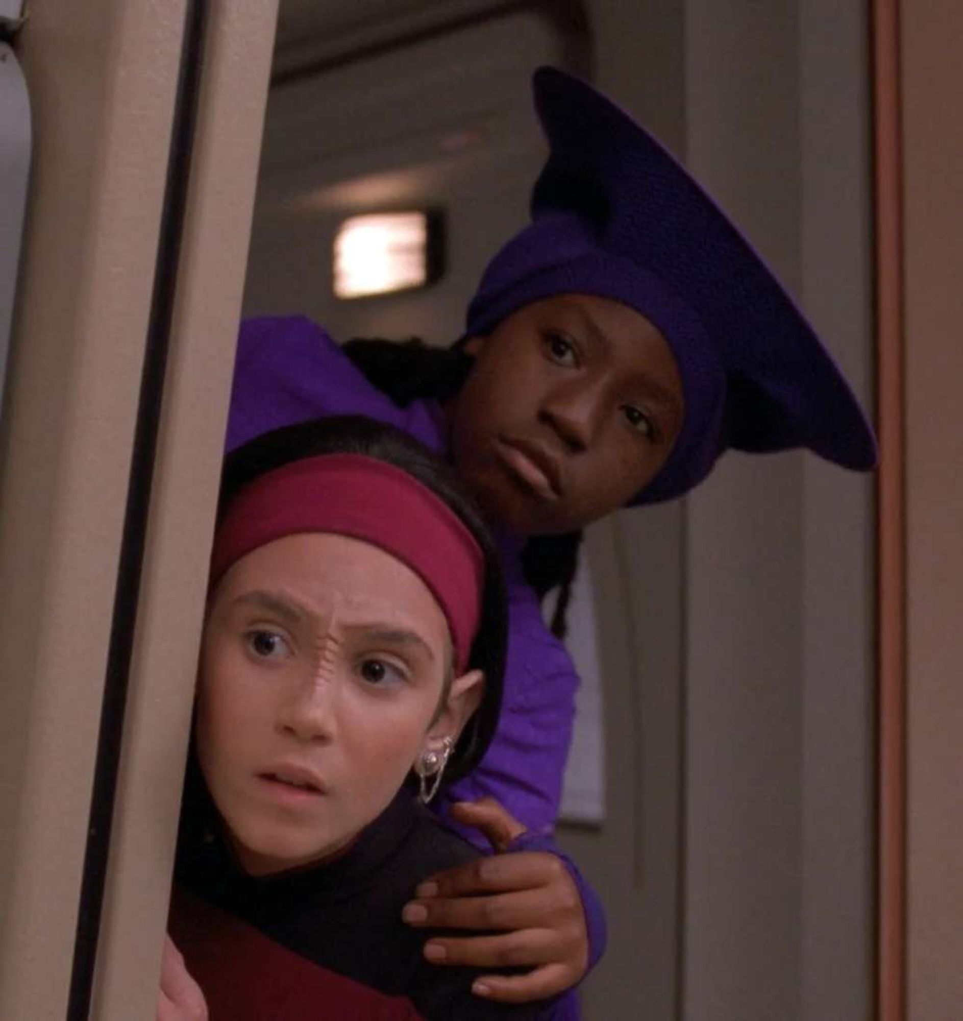 Young Guinan and Ro peeking around a corridor corner on the Enterprise.