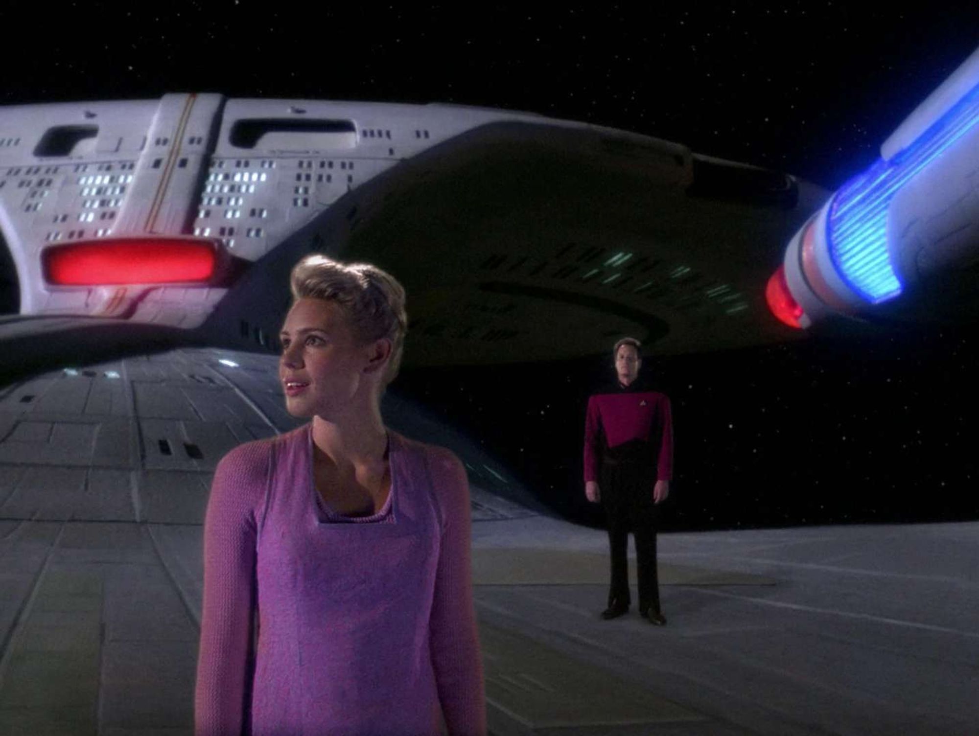 A blonde young woman on the hull of the Enterprise, marvelling at the space around her. Q stands in a captain's uniform behind her.