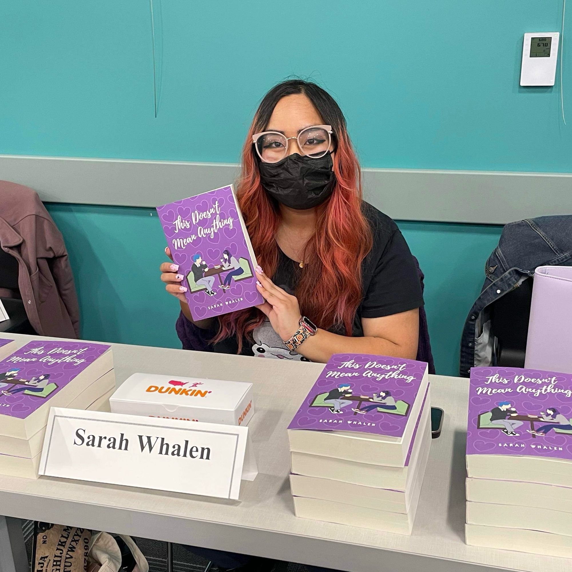 An Asian woman with long pink hair wearing a black mask holds up a paperback of THIS DOESN’T MEAN ANYTHING by Sarah Whalen. There are stacks of the paperbacks and a nameplate that says Sarah Whalen on the table she is sitting at.