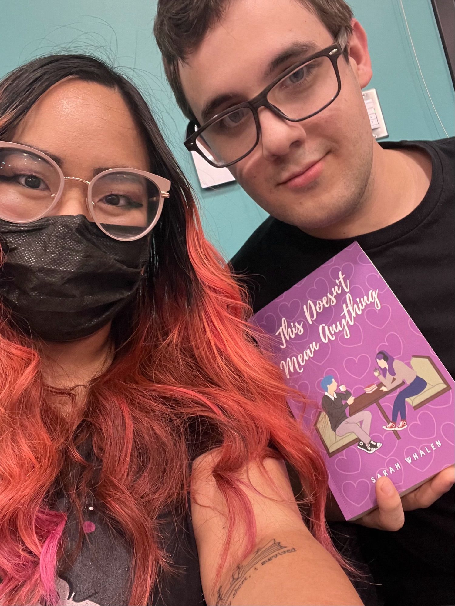 An Asian woman with long pink hair wearing a black mask next to a Caucasian man with dark hair. He is holding up a paperback of This Doesn’t Mean Anything by Sarah Whalen.