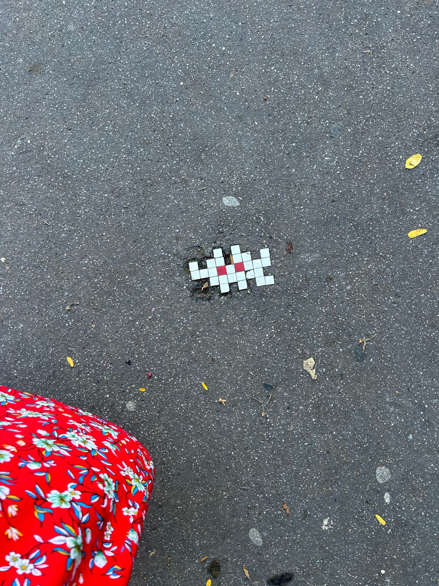 Space invader en mosaïque blanche, ses yeux en rouge, dans un trou dans le bitume. Il y a quelques feuilles jaunes éparses autour, et dans le coin gauche en bas, ma robe rouge à motif floral 