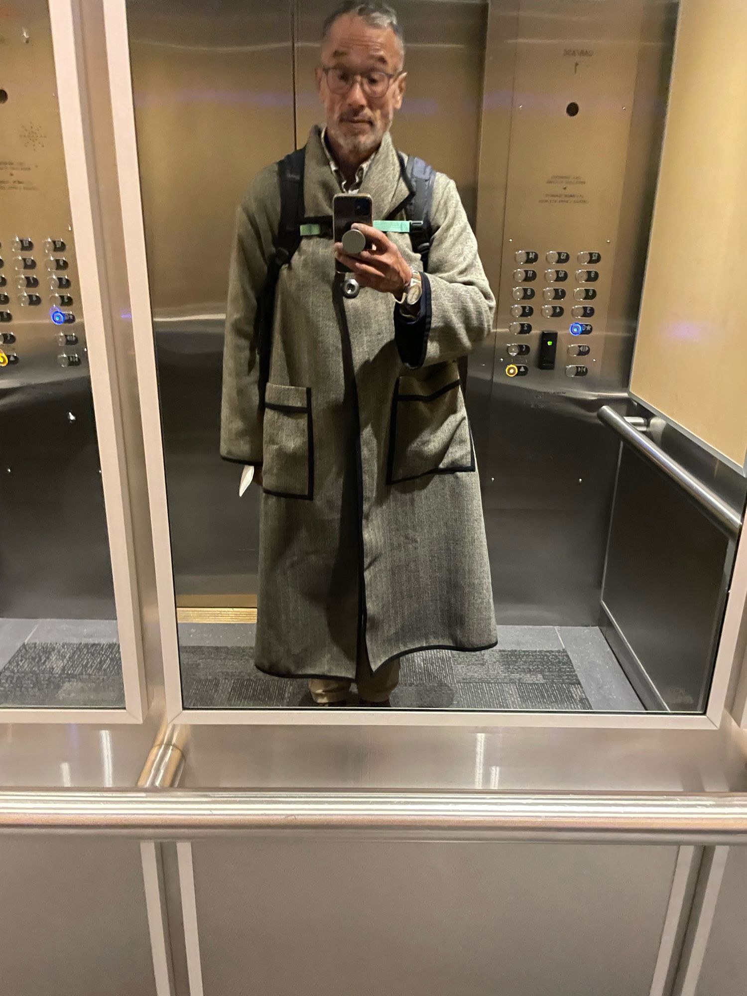 Taken in the work elevator wearing a long herringbone pattern coat with brown trim collar and sleeve. The coat is down to my shins with one brown button at the top so the coat opens up when walking. The sleeves are big oversized and two big pockets with a brown trim on the front of the coat.