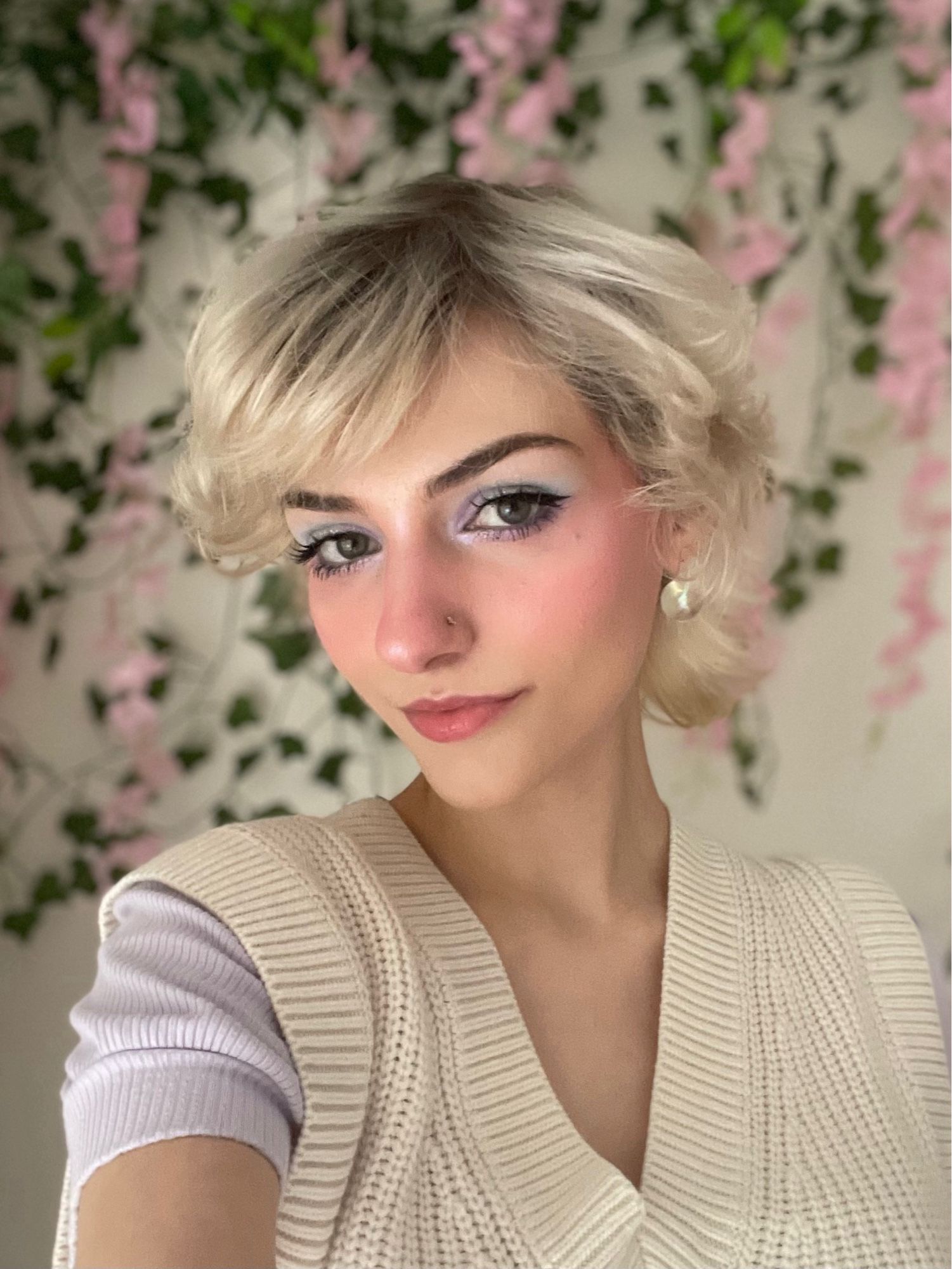 selfie of me in front of a wall of fake ivy and wisteria, my hair is kinda curly, and i have on blue/purple eyeshadow, my highlighter is slightly purple and im almost kinda smiling. my face is turned to the side and im wearing a purple blouse under a cream sweater vest.