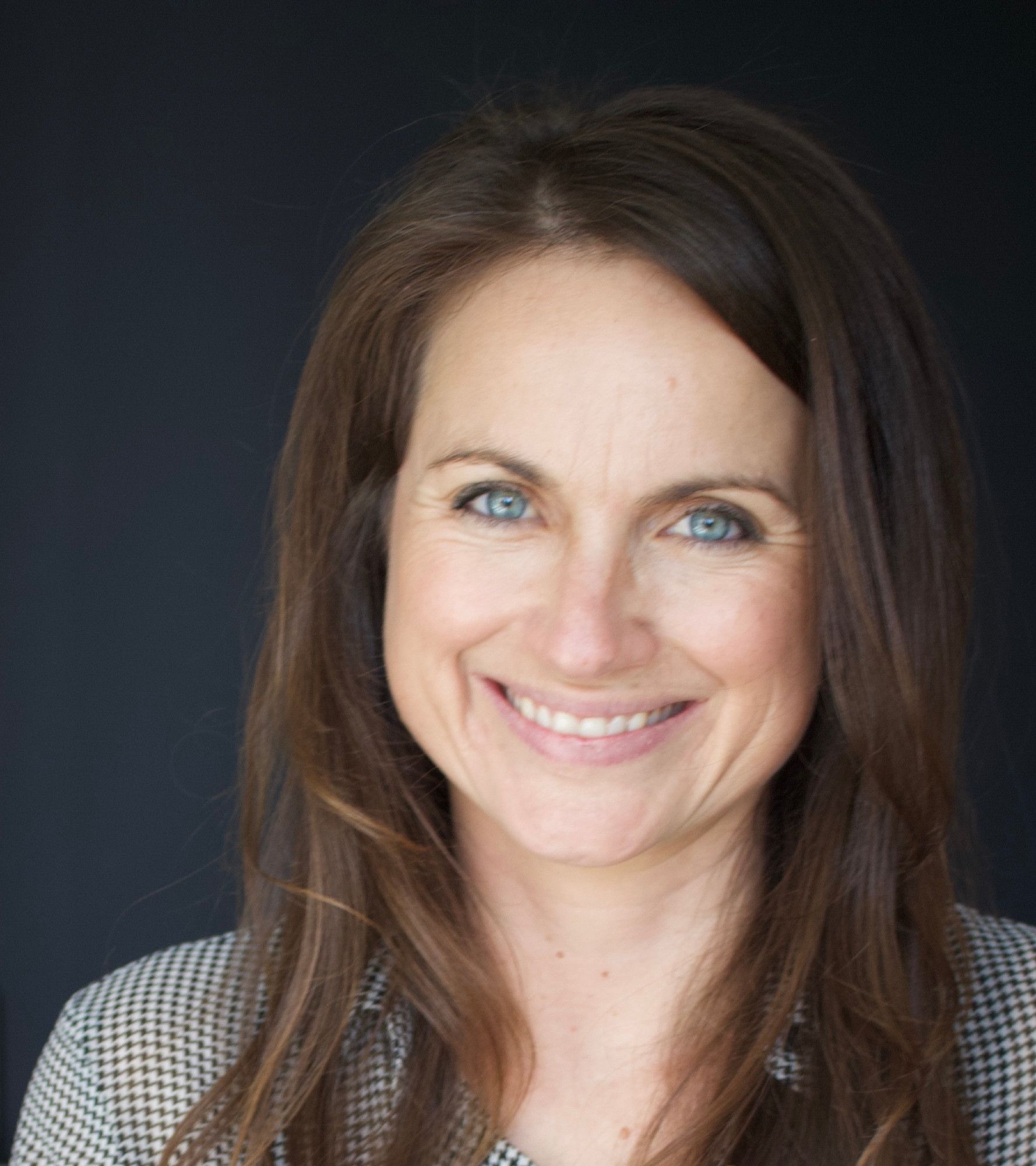 Headshot photo of the author quoted in this post. A white woman with long brown hair and blue eyes, wearing a black and white checked jacket.