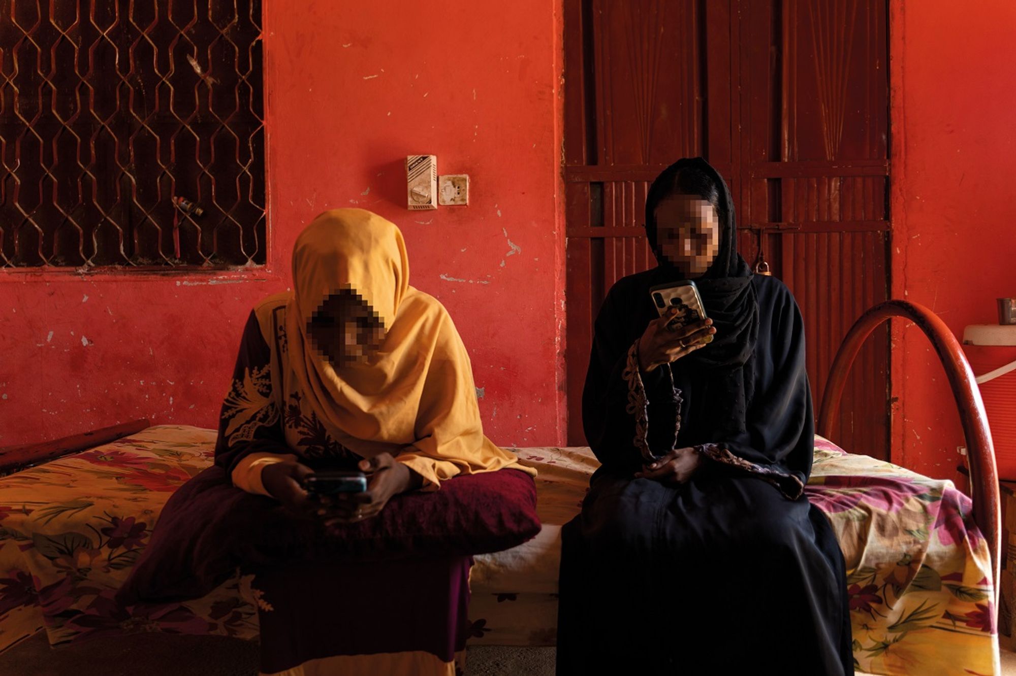 Photo credit: UNHCR/Ala Kheir

Caption: Internally displaced and living with relatives in White Nile State, Sudan. 

Description: Two young women (faces pixelated) sit on a yellow floral bedspread in room with bright reddish orange walls. One wears a black hijab and dress, and the other wears a bright yellow hijab and dress. They are both holding mobile phones and looking at the screens.