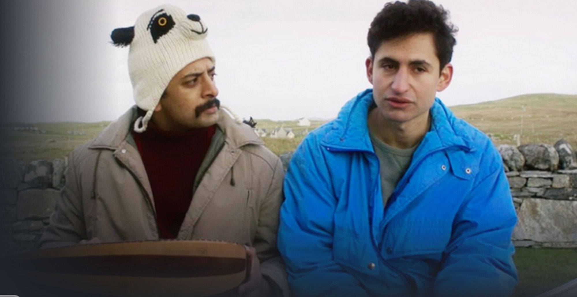 Two men sit in front of a low, old stone fence, some gentle hills behind the fence. One man is wearing a knitted panda bear winter hat and tan winter coat, open to reveal a maroon coloured jumper. A younger man sits next to him, with no hat and a blue winter coat.