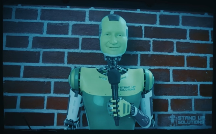 A green robot with a very human-looking face holding a microphone in front of a brick wall; it is KENN the "AI comedian" from Conner O'Malley's Stand Up Solutions comedy special.