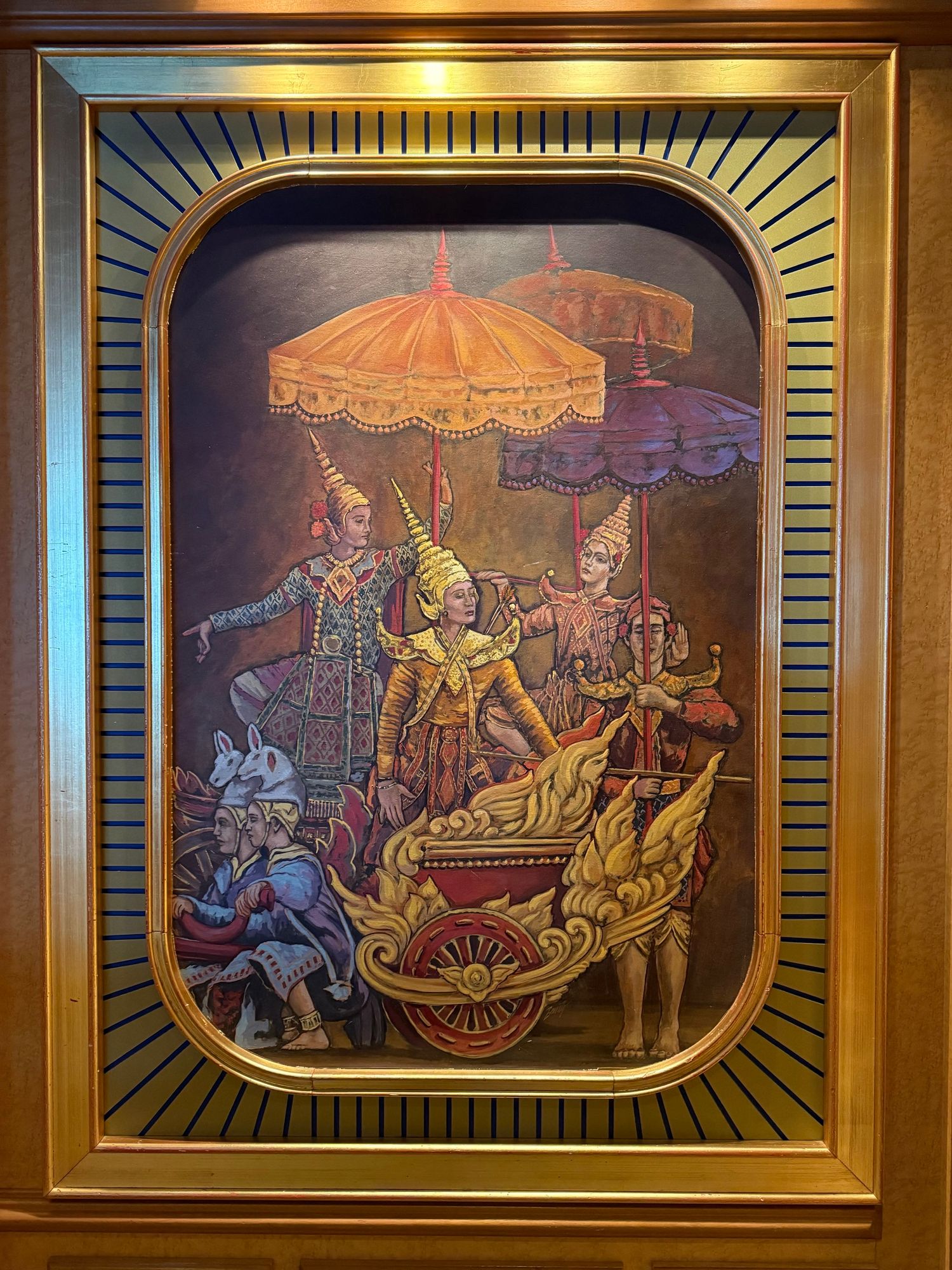 A framed painting depicting a traditional scene with several figures in ornate costumes. Prominent elements include golden chariots, elaborate umbrellas, and figures possibly representing historical or cultural characters, set against a richly textured background.