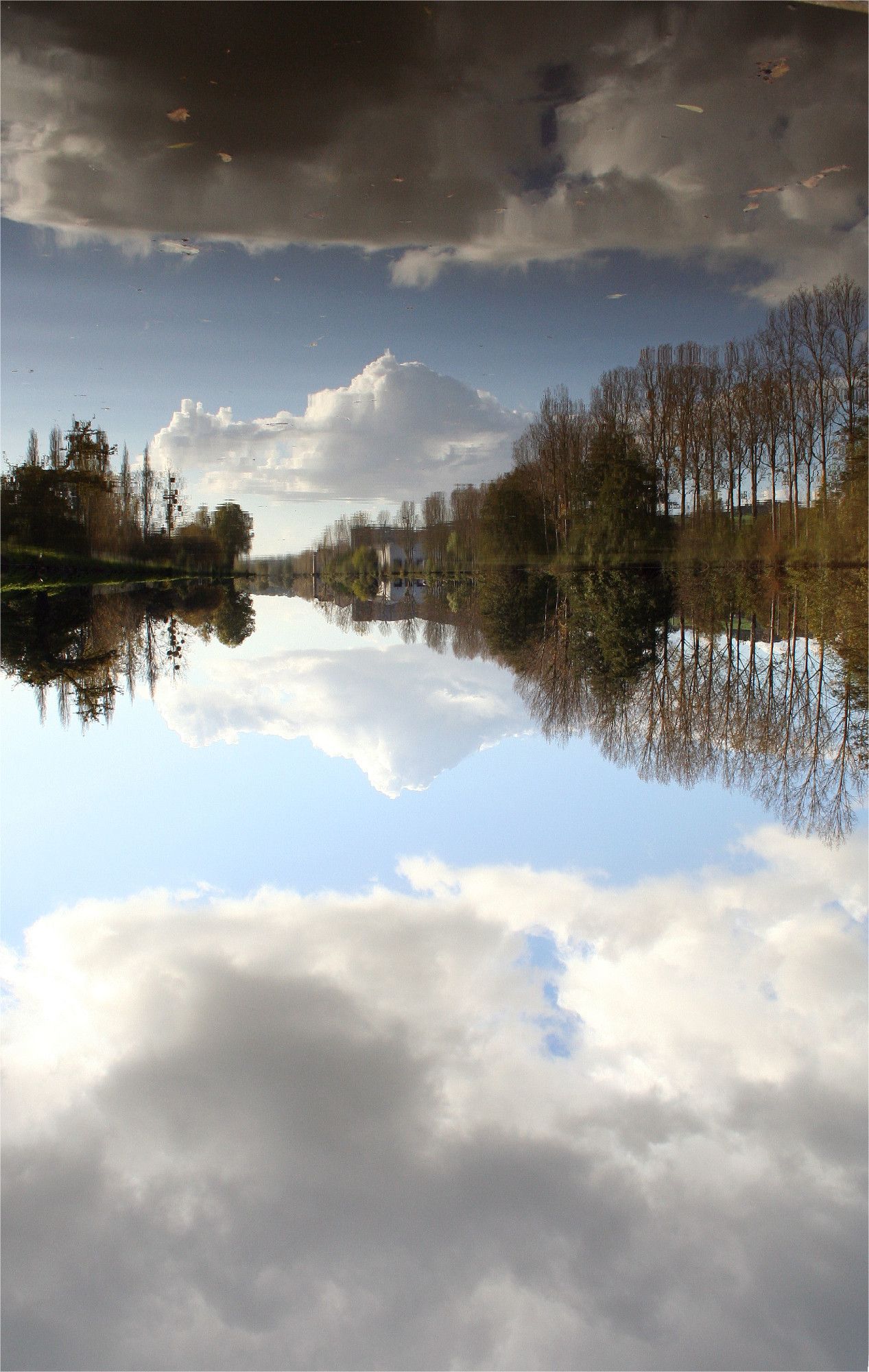 un paysage où les arbres et le ciel nuageux se reflètent parfaitement dans une étendue d'eau parfaitement calme, créant une symétrie. le ciel est bleu, nuageux et le ton des arbres révèlent l'approche de l'automne. Photo prise au milieu du pont qui surmonte le cours d'eau (le Semnon), Faite avec mon eos, cette photo est l'une de mes préférées.