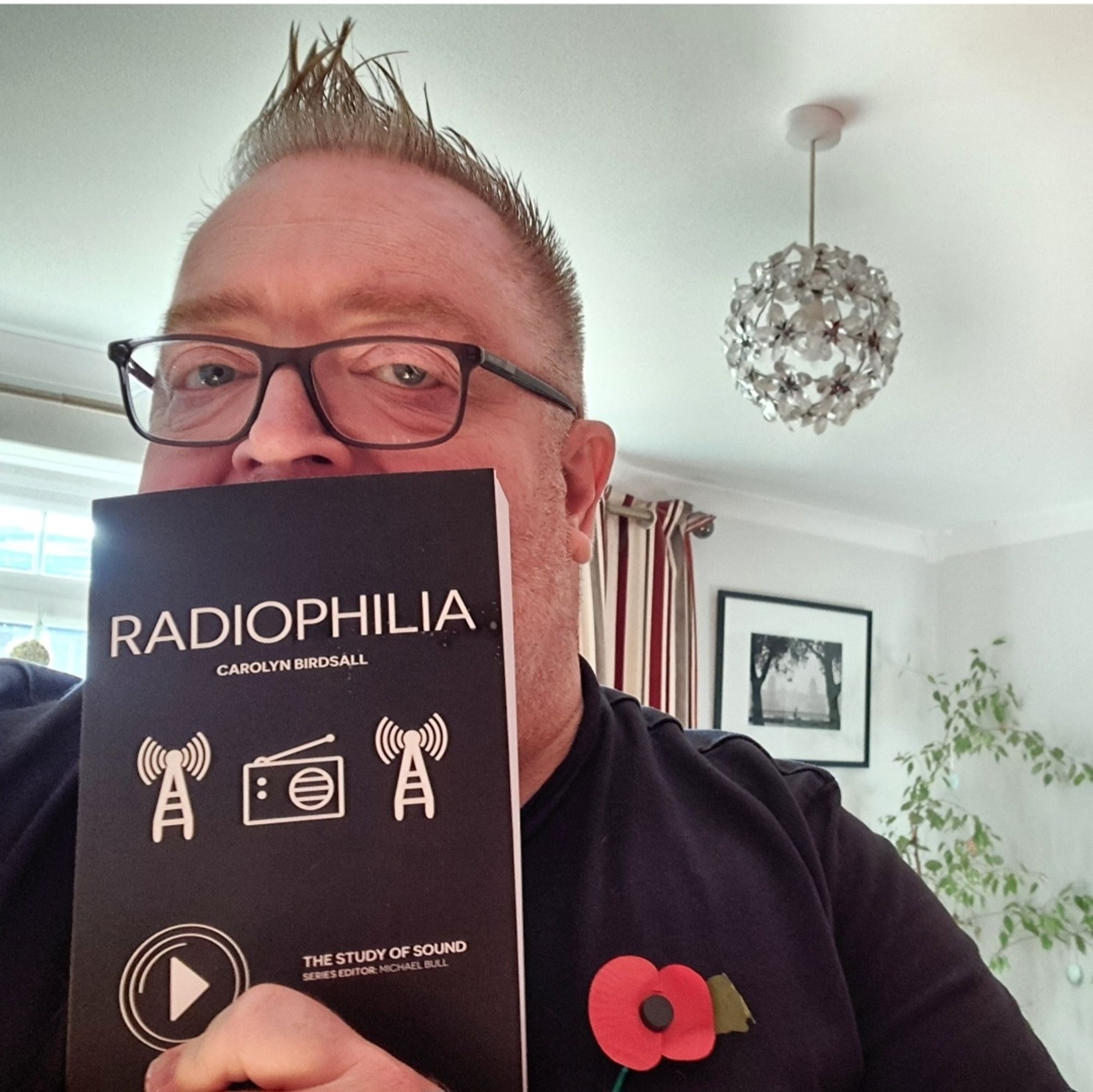 Me holding a copy of a book called by Radiophilia by Carolyn Birdsall, up in front of my face, obscuring my mouth. I'm wearing a poppy, glasses and a black t-shirt. You can see parts of my lounge in the background including a tall plant, a framed photo on the wall, and a glittery lampshade over my shoulder.