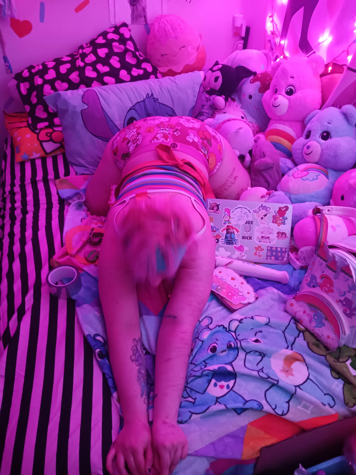 angel alog in a miami morty shift, dressed in neon with bleached hair. kneeling prone position on his bed. he has pink and black striped bed sheets. angel is face down, arching his back and sticking up his ass like a slut. he is surrounded by care bears, squishmallows, his care bear backpack, holographic duct tape, dab containers and a nectar collector, and his hitachi magic wand. his room is lit by pink string lights.