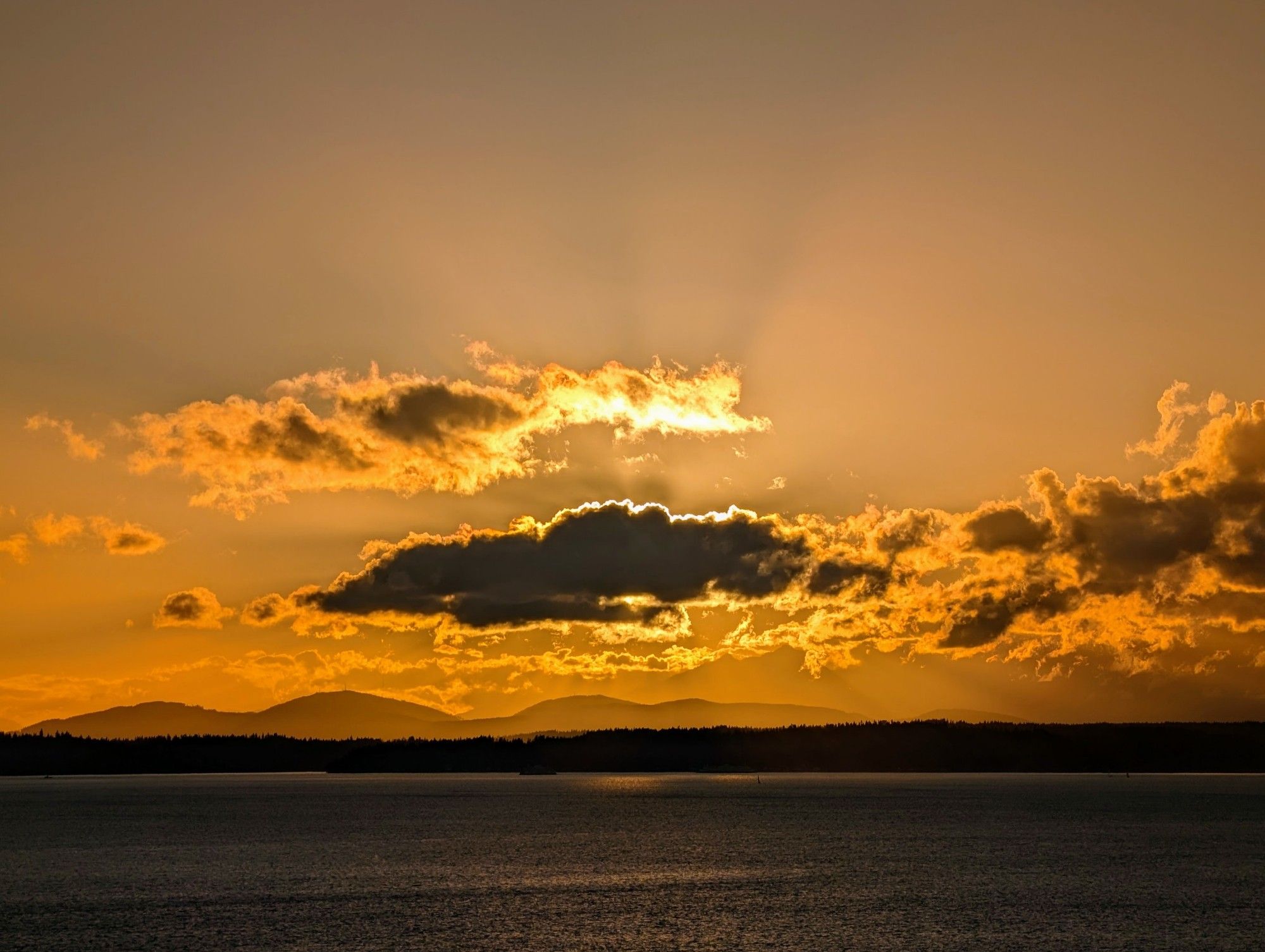 the setting sun obscured by a cluster of fluffy clouds with bright rays of light outlining the clouds, all over the Seattle waterfront. mountains and the small islands in the distance.