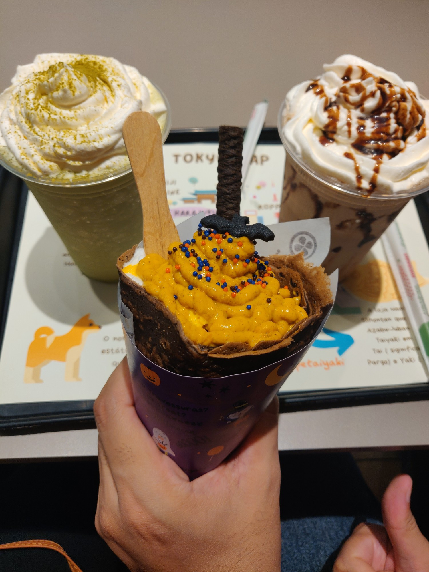 Duas bebidas de frapê ao fundo, uma de matcha e outra de mocha.
Na frente, temos um crepe de halloween com um pequeno morcego de decoração.