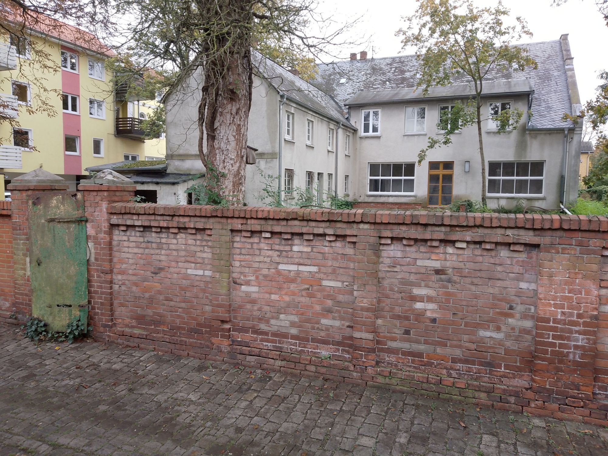 Im Vordergrund eine ca. 2 Meter hohe Mauer.
Dahinter das oben beschriebene Haus.
Auf der Rückseite also sind zwei sehr große Fenster und eine Holztür neu eingebaut.