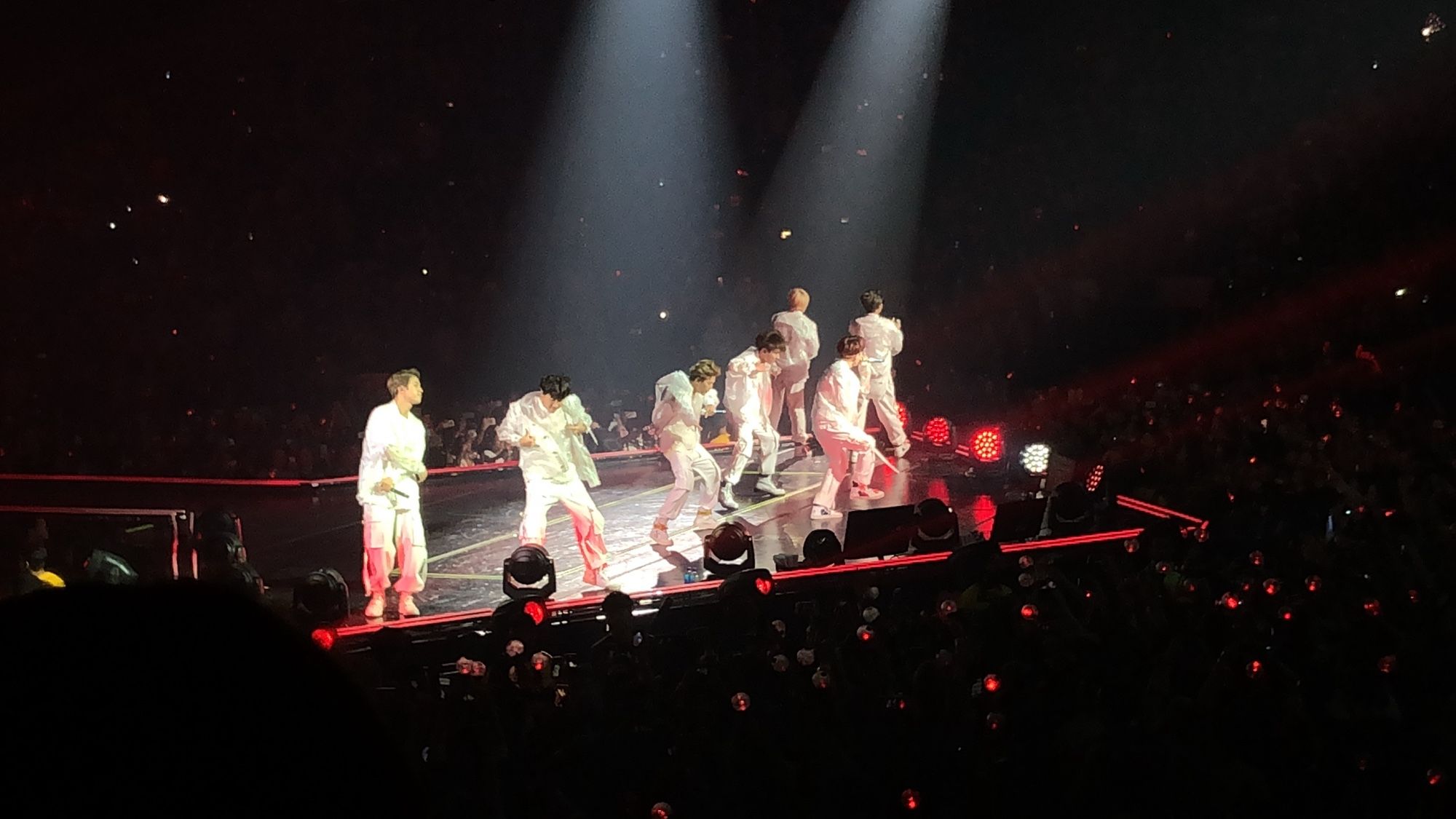 BTS on stage performing. The band members are all dressed in white with lights shining in them while the crowd is lit in red