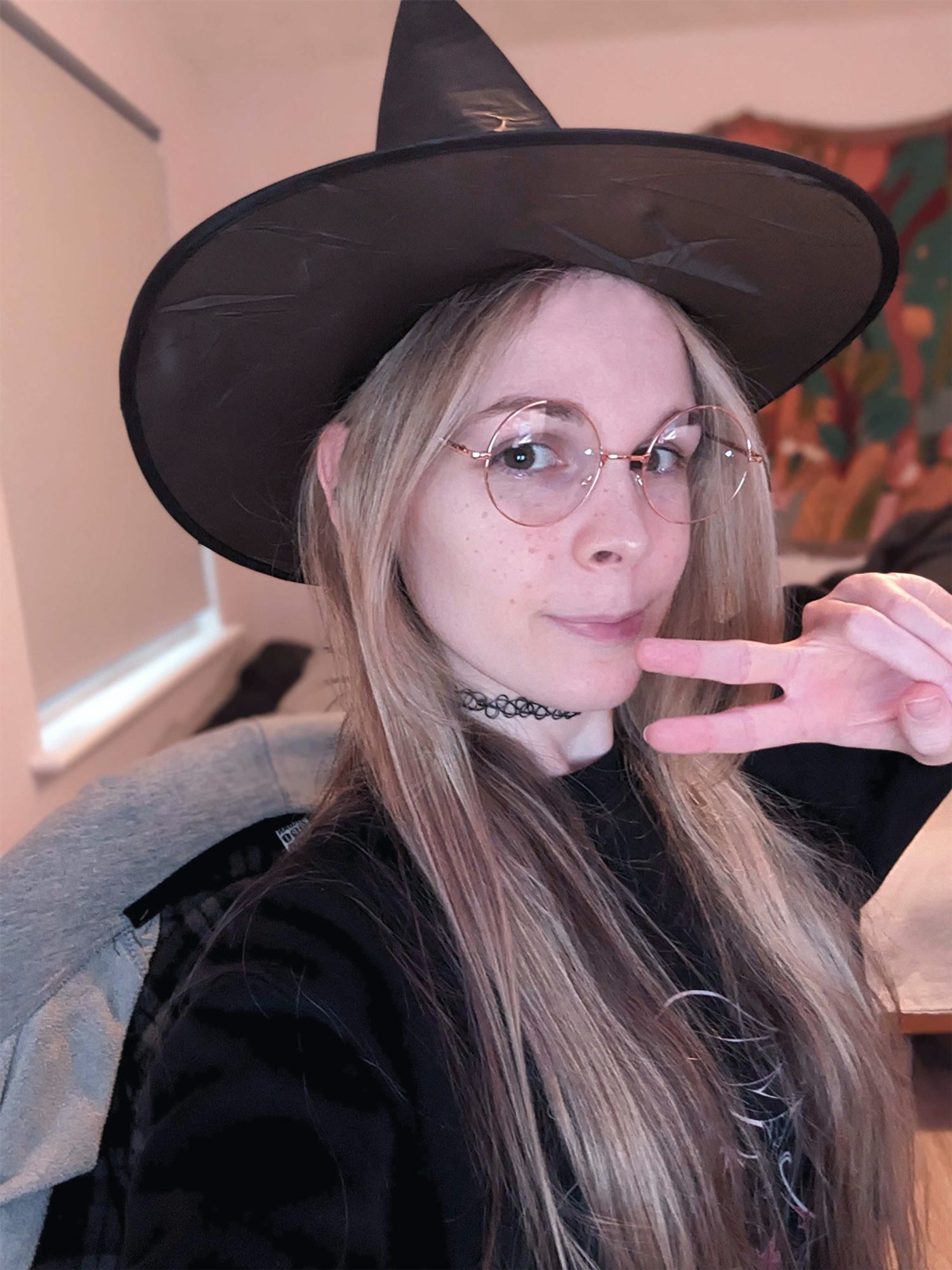 Charlotte wearing a witches' hat, round glasses, a choker and a black sweater. She's doing a little peace sign like a homo.