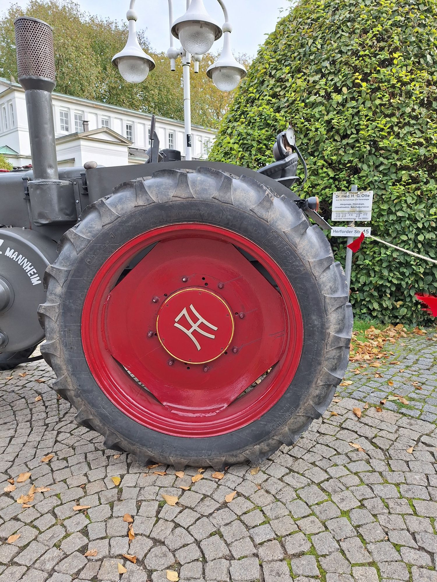 Hochformat, Farbe.  Foto zeigt das riesige Hinterrad des Lanz Bulldog  mit dem grobstolligen Reifen und der roten Felge auf der das typische Lanz-Symbol mit dem großen H in dem mittig noch ein L steht.