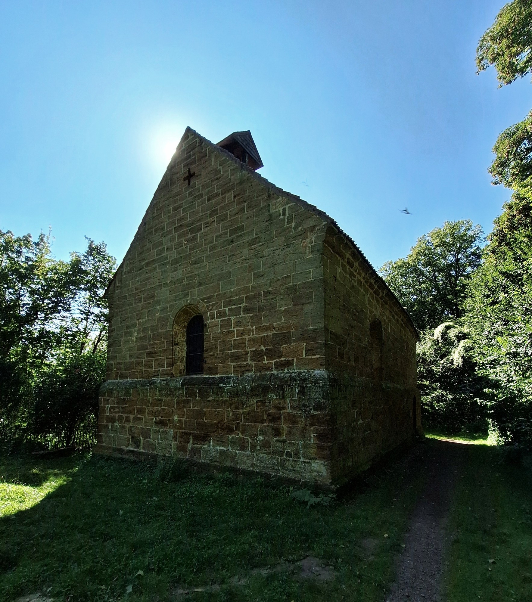 Ein Kirchengeebäude aus Kalksandstein im Gegenlicht auf einer grünen Wiese umgeben von Sträuchern und Bäumen.