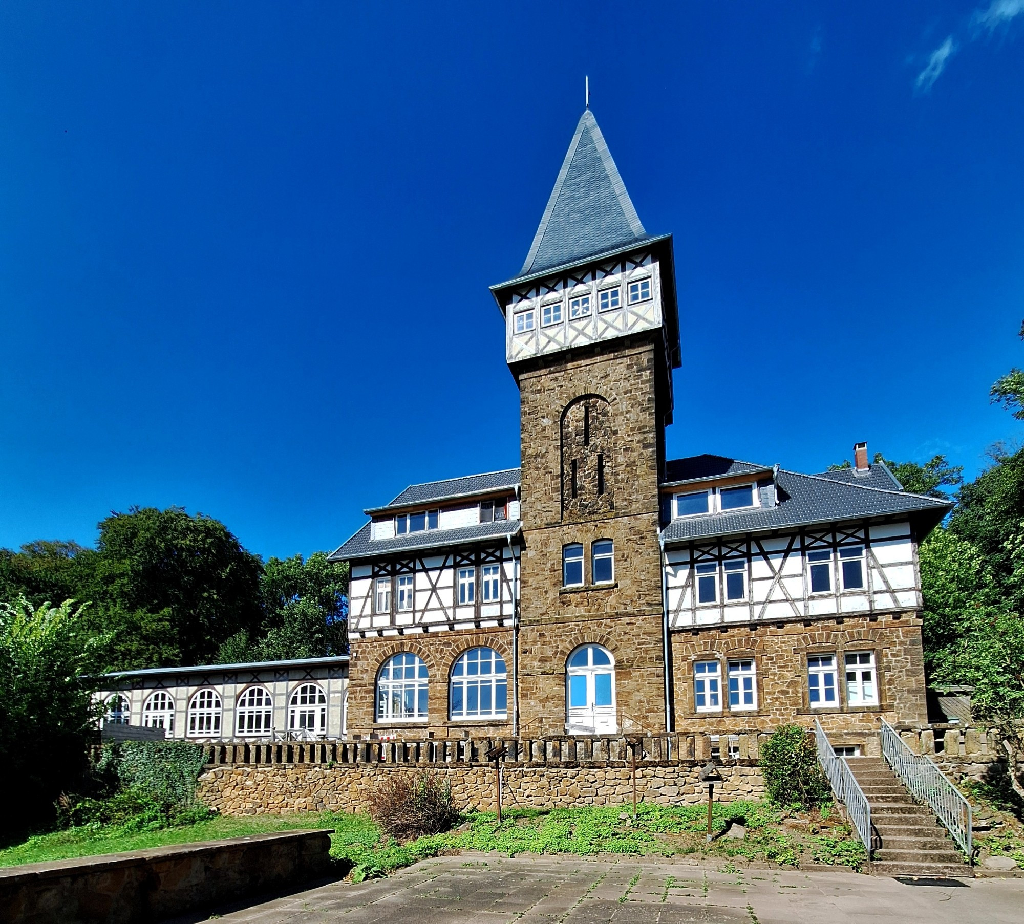 Hochformat, Farbe. Ein altes Gebäude aus Kalksandstein  mit Fachwerk und in der Mitte ein hoher Turm mit spitzem  Dach. Davor ein kleiner Platz und ein paar grüne Sträucher. Der Himmel ist wolkenlos blau.