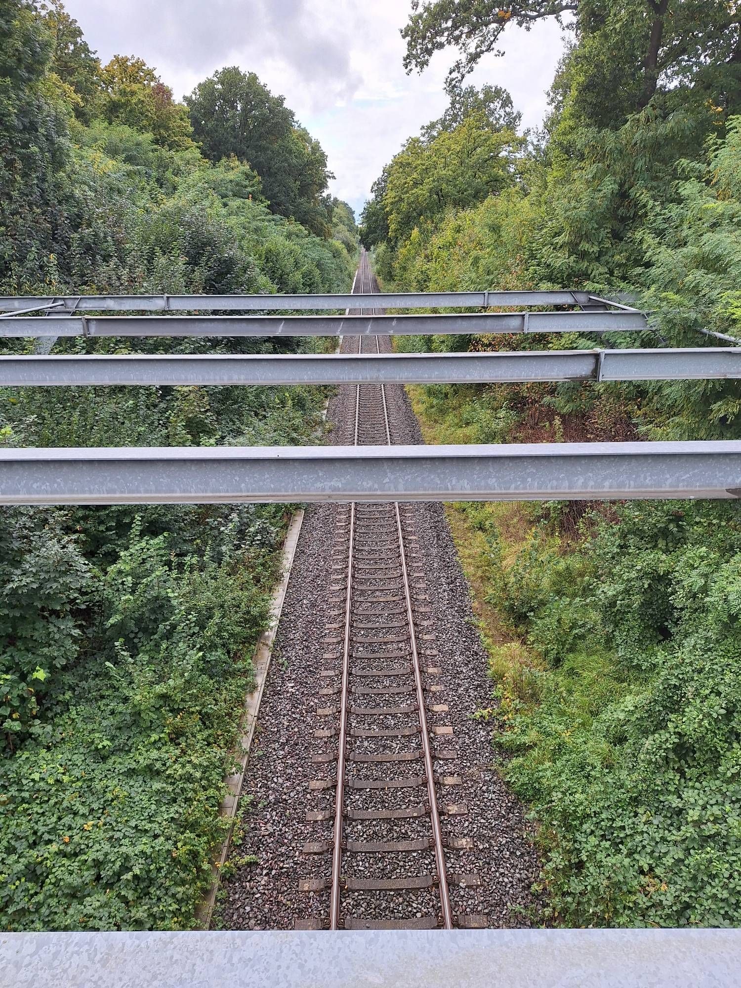 Hochformat, Farbe, man sieht graue Stahlträger die quer durch das Bild verlaufen und eine Eisenbahnstrecke überspannen. Rechts und links des Bahndammes ist eine grüne Böschung. Der Himmel am Horizont ist regengrau bewölkt.