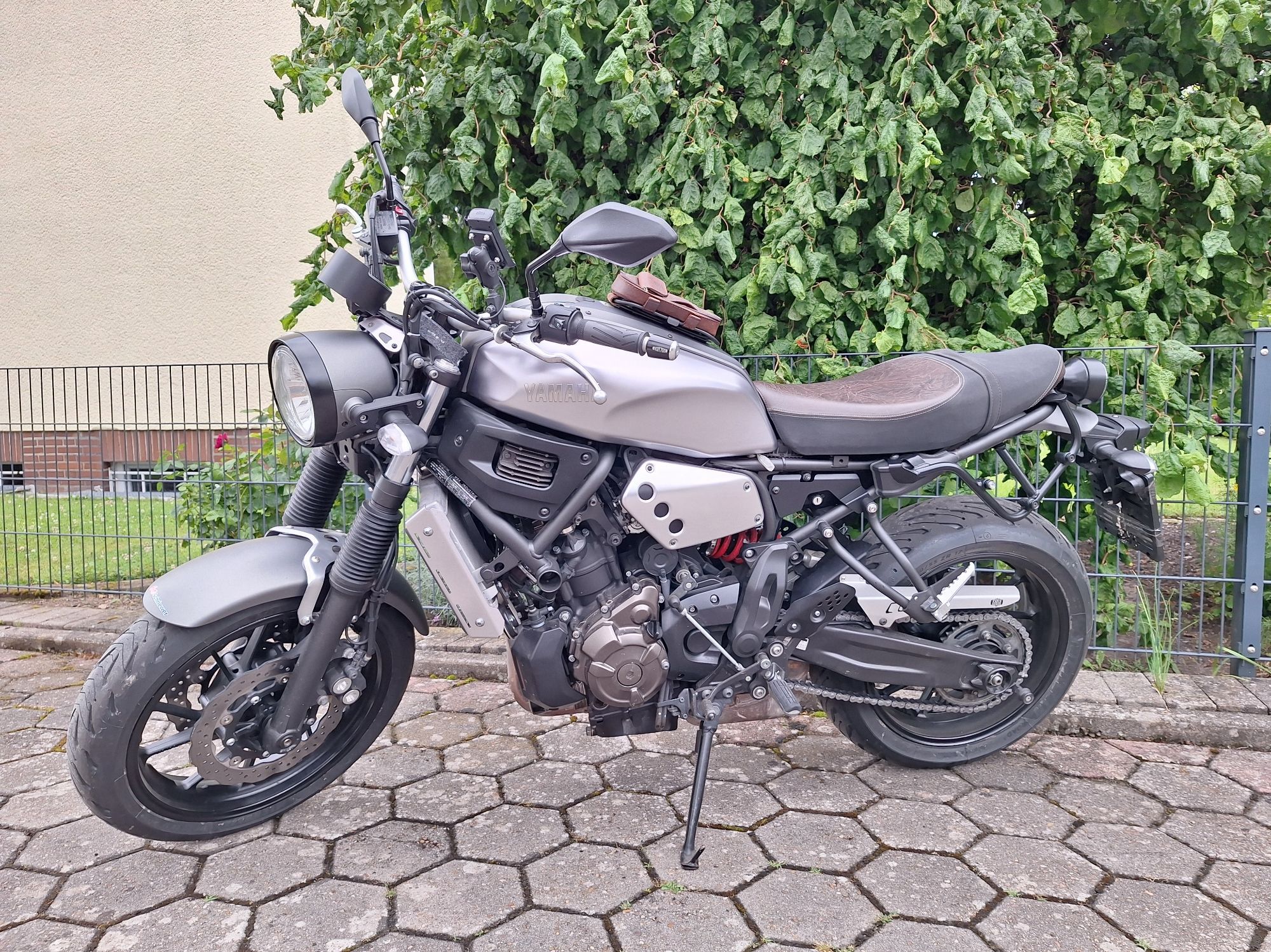 Ein silbernes Motorrad auf unserem Hof vor einem grünen Strauch und der gelb angemalten Hauswand der Nachbarn.
Das Motorrad hat einen schwarzgrauen Runden Scheinwerfer, einen schwarzen Motor und eine Schwarze Sitzbank die auf der Sitzfläche braun abgesetzt ist. Auf dem Silbernen Tank ist ein kleiner Gepäckträger mit einer braunen Tasche.
Das Motorrad besitzt keine Verkleidung. Ein sog. Nakedbike.