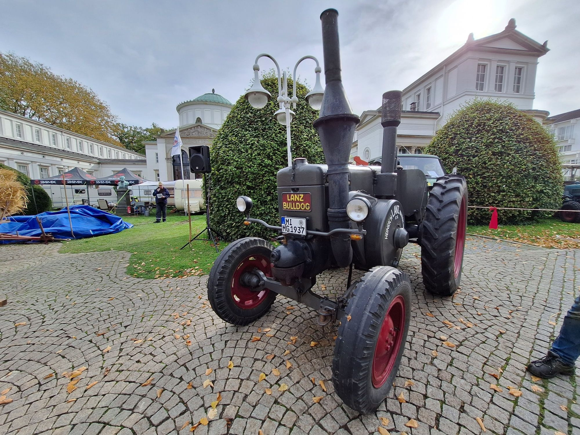 Querformat, Farbe.  Foto zeigt einen alten Traktor in schwarz. Es ist ein Lanz Bulldog mit mächtigen Auspuff mit dickem Funkenkobel. Auf dem Ansaugrohr sitzt ein Ansaugkäfig. Im Hintergrund sieht man ein altes, weißes Gebäude aus dem Kurpark, das Badehaus I.