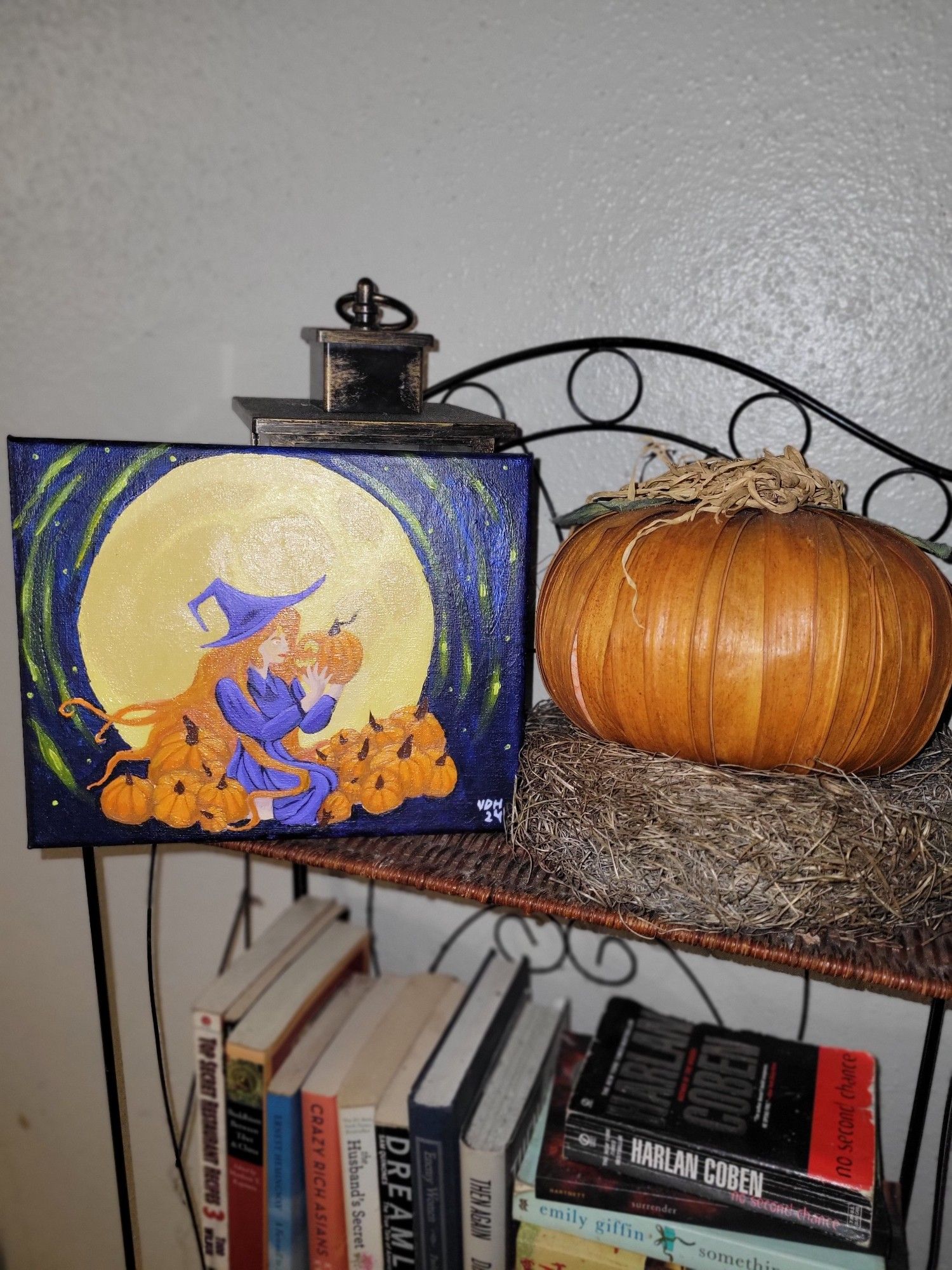 A finished painting of a witch with pumpkins in front of a large glowing moon sits next to a fake pumpkin on a wicker bookshelf.