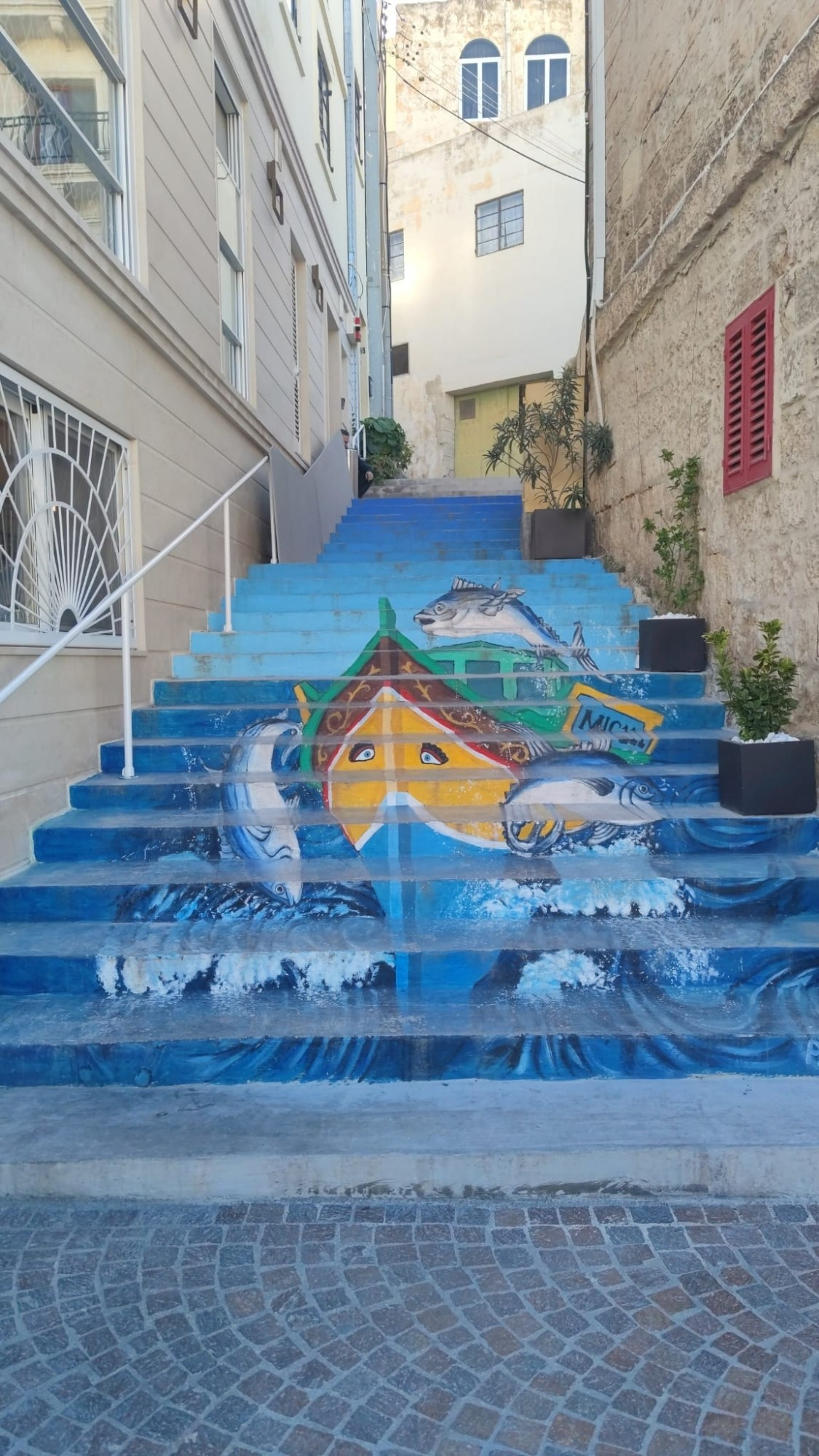 Eine Treppe in Mellieha, kleine Stadt auf Malta. Wenn man von unten nach oben schaut, erkennt man ein farbenfrohes Schiff, von Wellen umgeben. Links ist ein Treppengeländer, rechts sind Blumenkübel aufgestellt. Es dominieren blaue Farben.