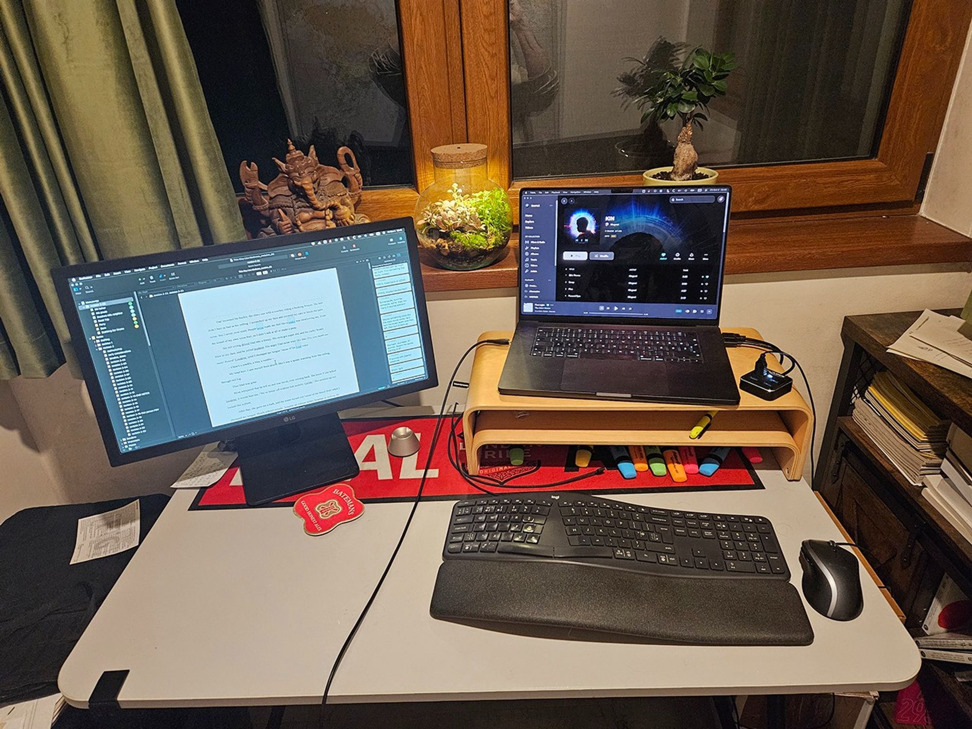 My writing desk with Scrivener open on the left monitor, and Tidal Music open on the Mac, playing the band Mogwai. Ganesh watches over me, along with some plants.