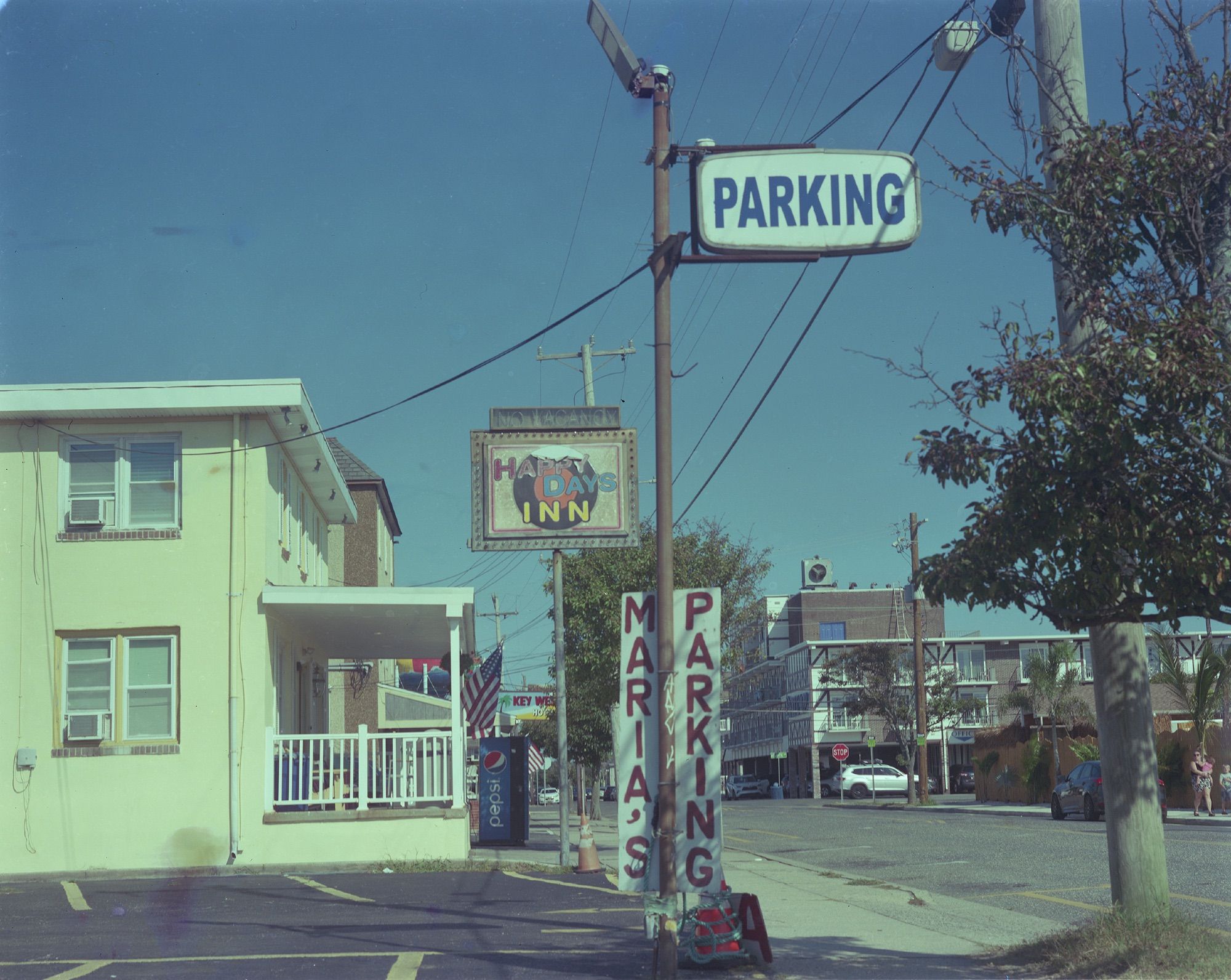 A motel stands behind a parking lot. The parking lot has a sign that says “Parking” at the top and “Maria’s Parking” hand painted attached to either side of the sign pole. The motel has a sign that says “Happy Days Inn” over an illustration of an LP.