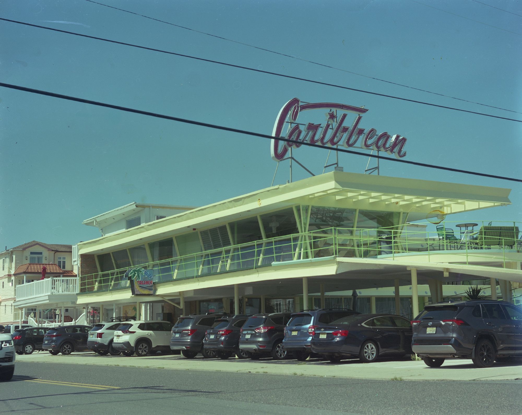 A mid-century modern hotel with a large neon sign on the top that says “Caribbean” in script. Electric wires cross the scene, and the neon sign is largely between the wires. The parking lot is full.