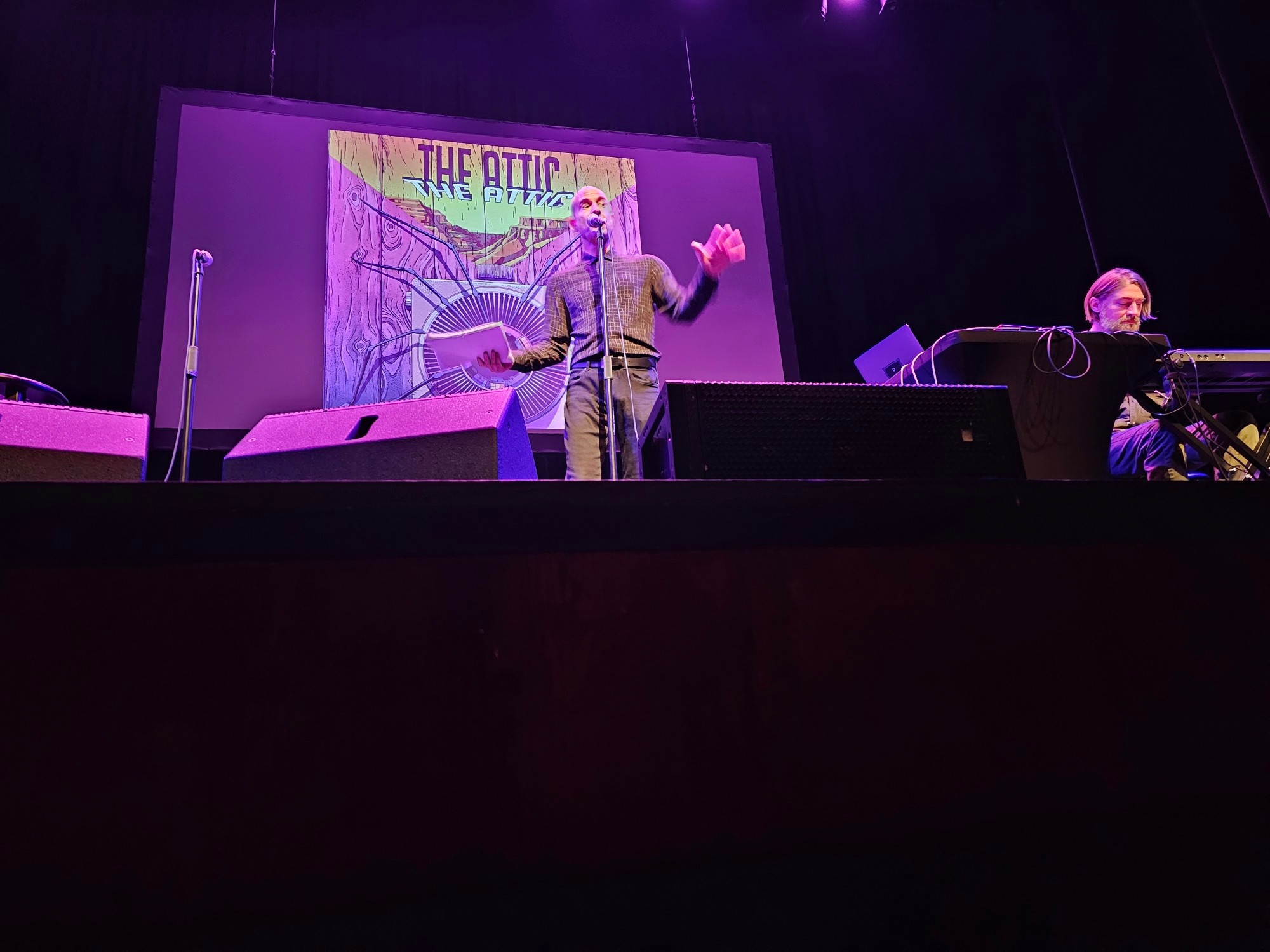 The actor Cecil Baldwin stands on a stage with purple lighting, a slide stating "The Attic" behind him. To his left in a small soundbooth sits musical composer Disparition.