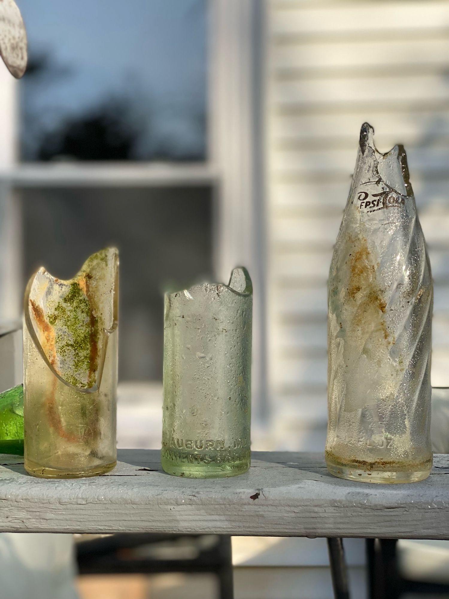 3 glass bottles with stains from the ocean are on a gray porch with a window in the background.