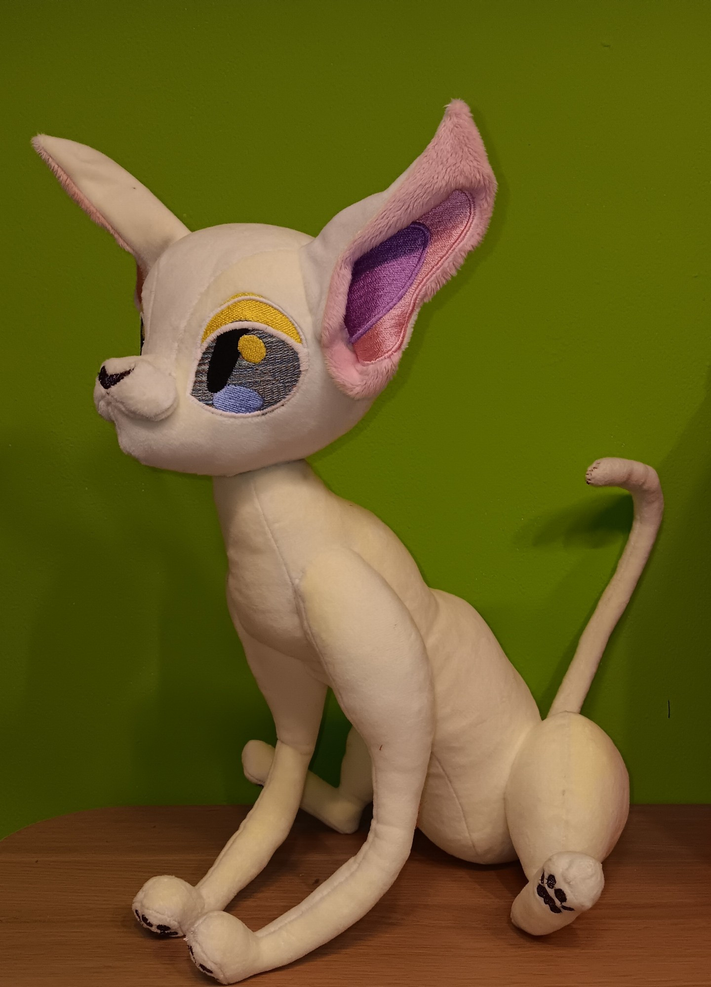 A plush of a sphynx cat sitting in Front of a green wall. Her skin is White with beige/Creme coloured, her nose and paw pads are Black with glitter. Her eyes have a bluish color gradient with yellow eye lids