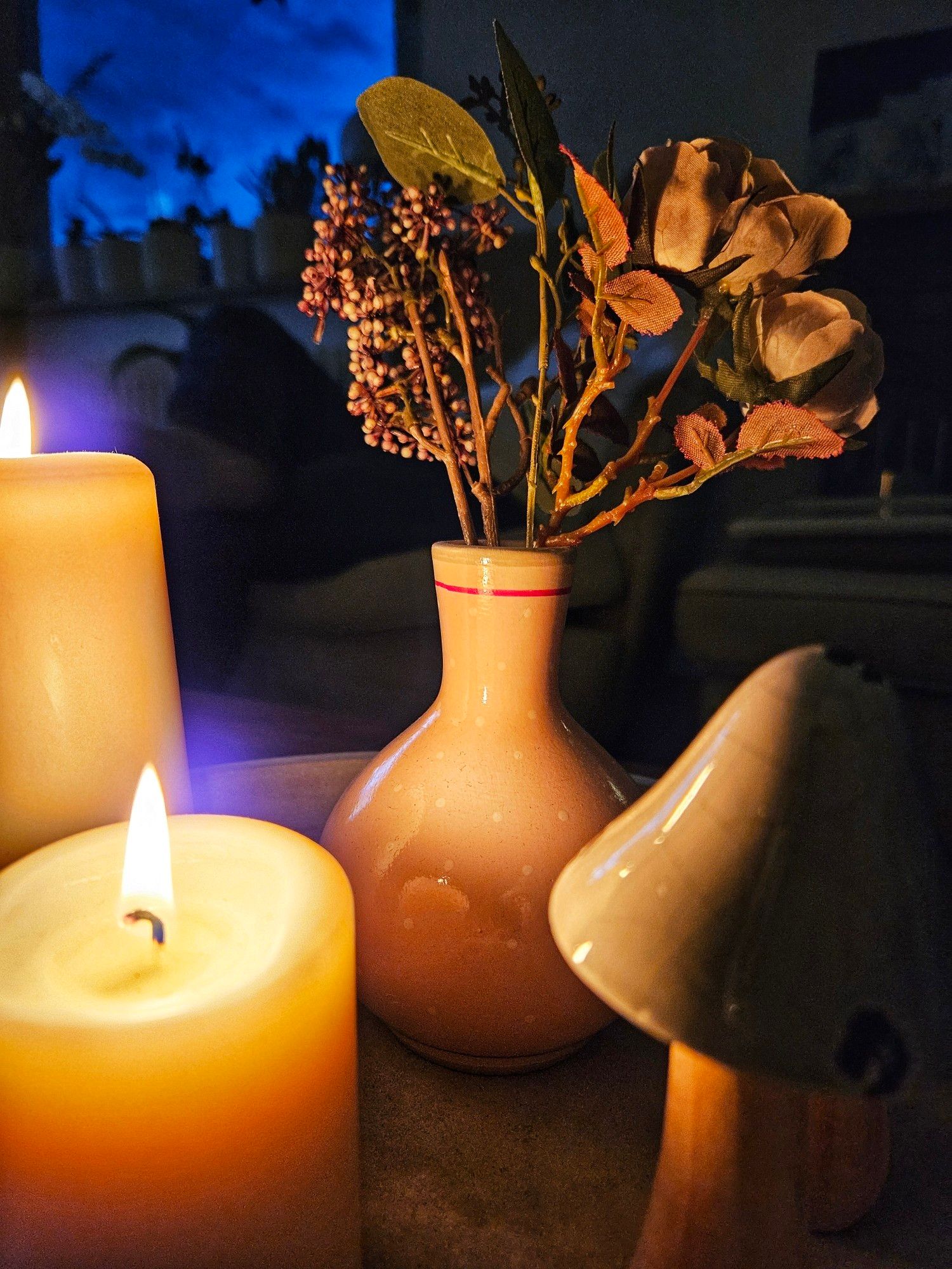 Zwei Kerzen links, ein Steingut-Pilz rechts, im Hintergrund eine Vase mit Trocken Blümchen, alles im warmen Kerzenlicht. Dahinter ein Fenster mit dunkelblauem Himmel-Hintergrund.