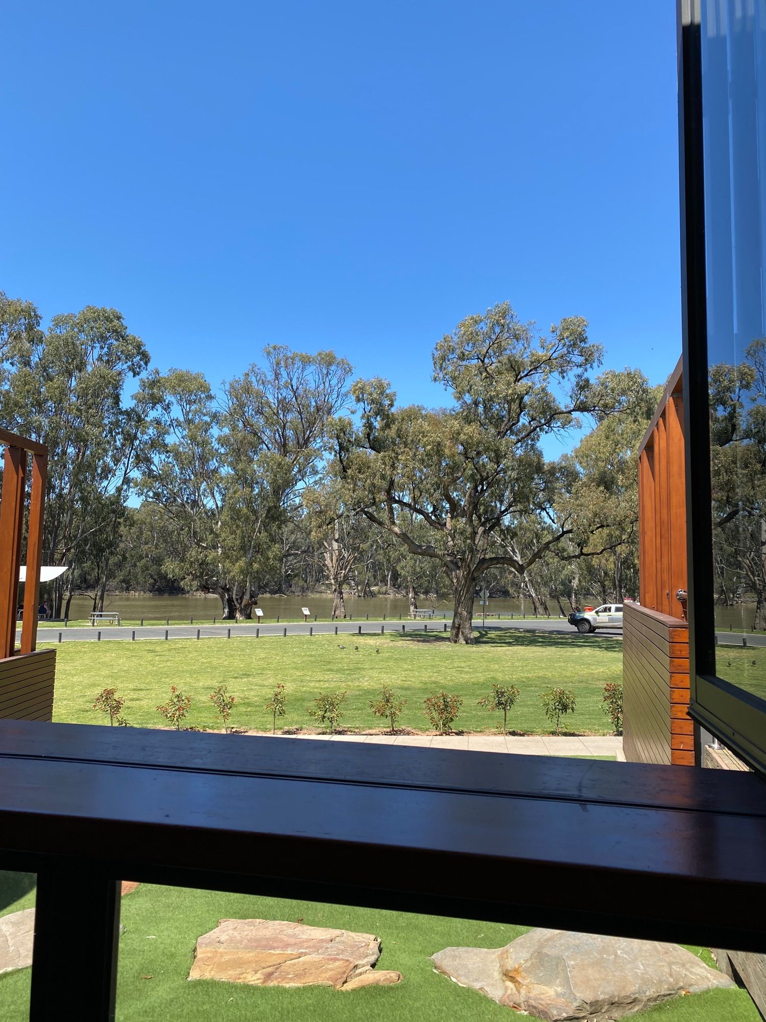 View from balcony overlooking grass with trees lining river bank in distance. Bright blue sunny sky overhead.