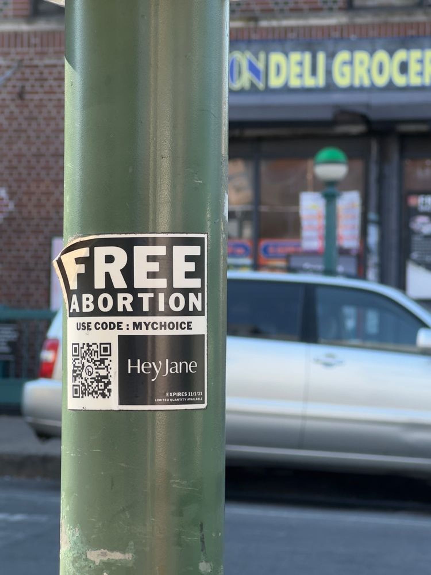 A imagem mostra um poste de cor verde com um adesivo colado que anuncia “Free Abortion” (aborto gratuito) com um código promocional “MYCHOICE”. Abaixo do texto, há um QR code e o nome “Hey Jane” com uma data de expiração de 11/1/21. Ao fundo, é possível ver parte de uma loja de esquina chamada “Deli Grocery” e um carro estacionado na rua.