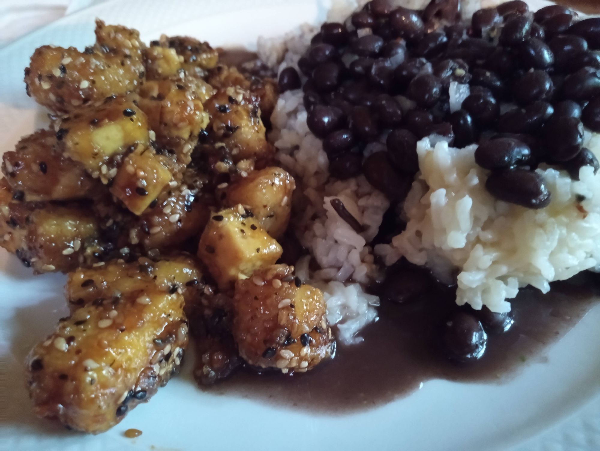 A white dinner plate with white rice that is topped with black beans (on the right side) and sauteed tofu in teriyaki sauce on the left side.