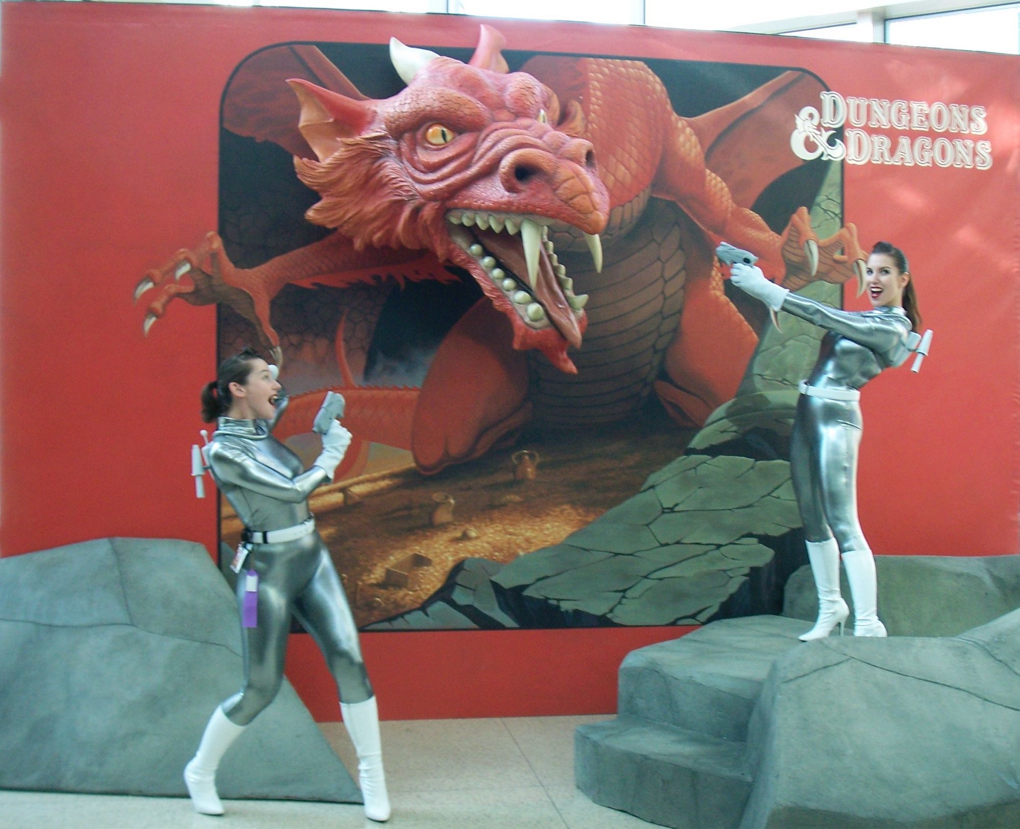 A photo of two women in 1950s style shiny silver form-fitting space suits posing next to a large stand-up display ad for Dungeons & Dragons featuring a red dragon. The red dragon’s head is a 3D physical element that appears to be emerging out of a cave. The women are pointing sci-fi pistols at the dragon, recreating an action scene as it would appear on the cover of a magazine or game product.