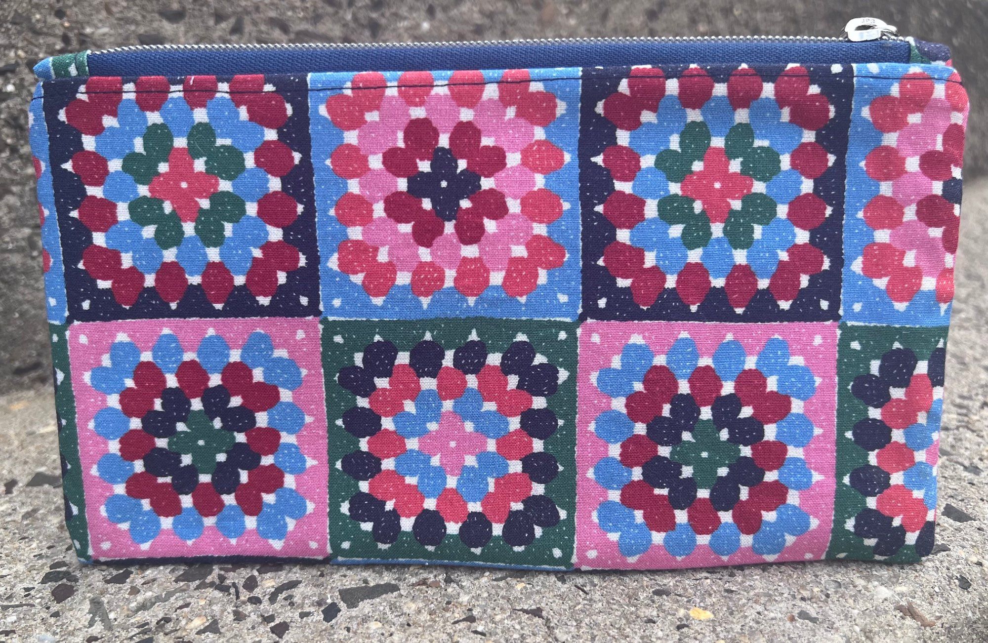 A sewn flat-zip pouch that measures 9"x5" and has granny squares printed on the fabric in a navy, pink, red, and green pattern.