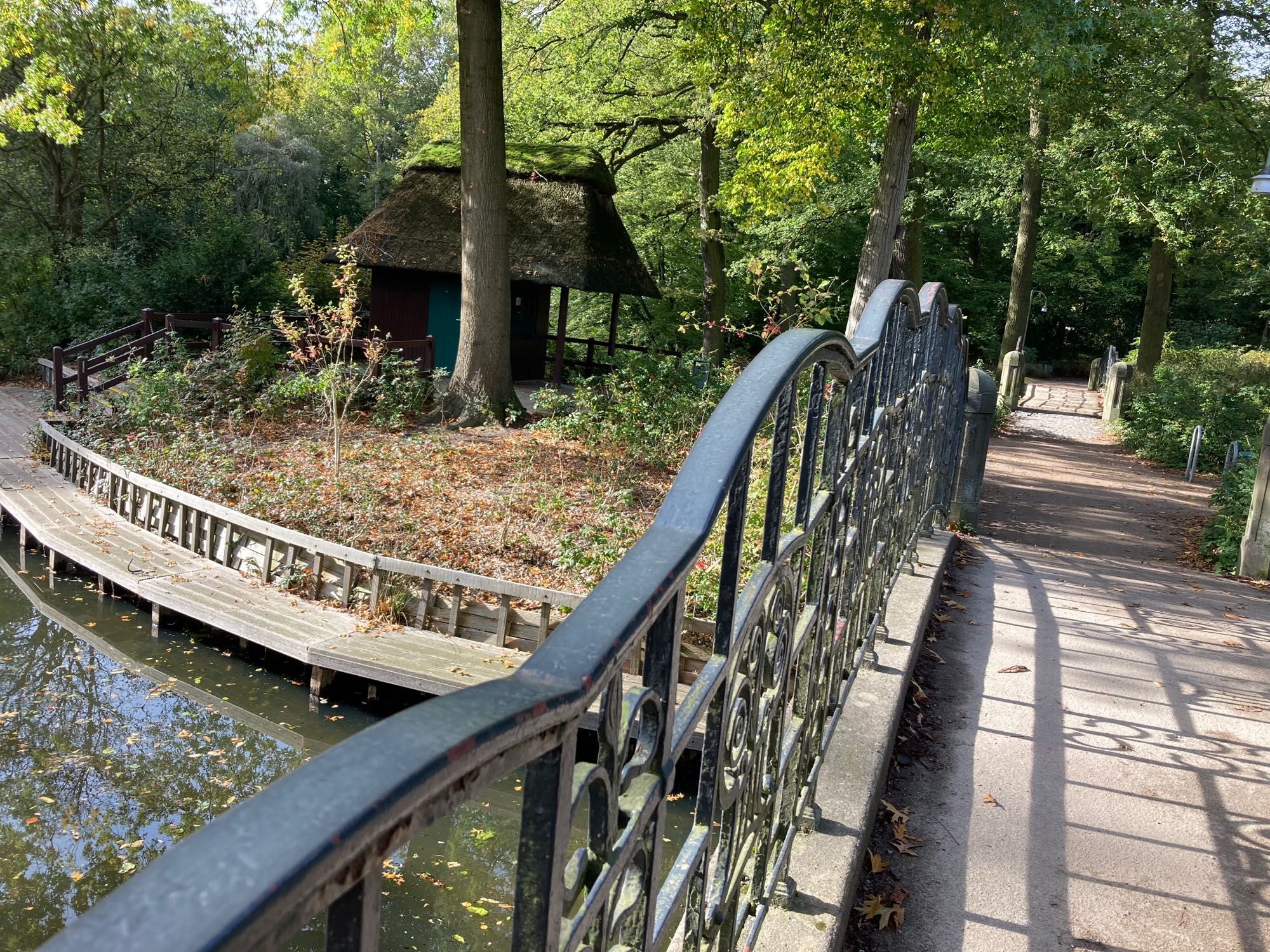 Das Foto zeigt eine wellenförmige Brücke mit geschwungenem Geländer. Links im Vordergrund etwas Wasser vom Emmasee. Ein halbrunder Steg begrenzt das Ufer nach hinten zu. Im Hintergrund stehen eine Hütte und Bäume.