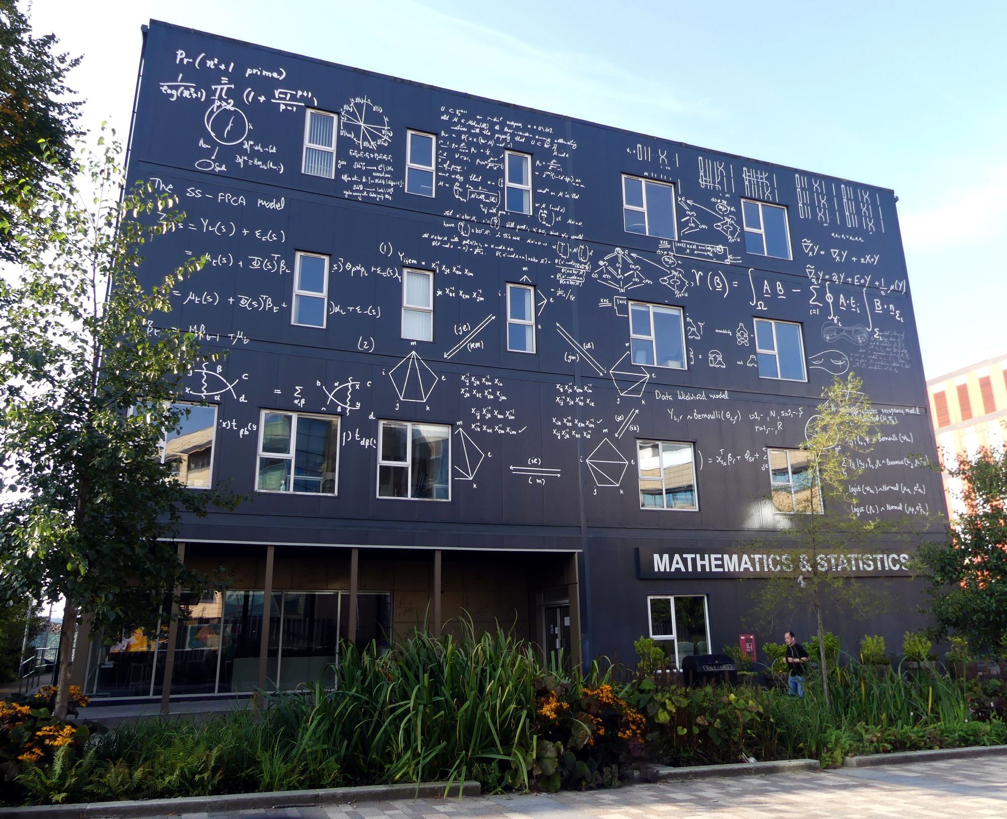 A boxy modern university building, labelled "Mathematics & Statistics", painted in a dark colour and decorated with chalkoard maths.