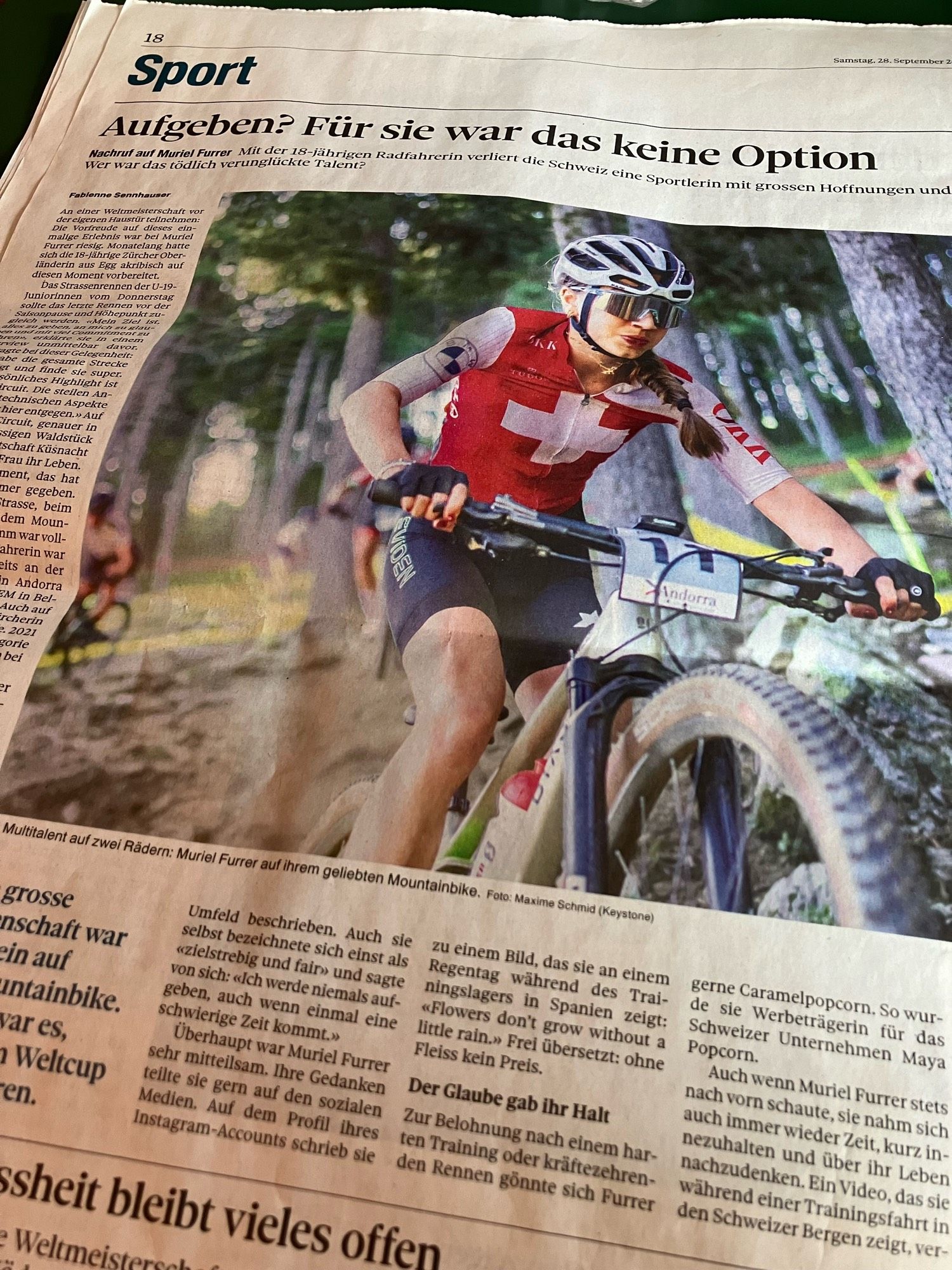 zeitungsausschnitt.
oben rechts, gross: sport
titel: Aufgeben? Für sie war das keine Option
lead: Nachruf auf Muriel Furrer [fett] Mit der 18-jährigen Radfahrerin verliert die Schweiz eine Sportlerin mit grossen Hoffnungen und Träumen. Er war das tödlich verunglückte Talent?
[grosses bild. junge frau mit rotem lippenstift im kurzarm-velo-dress von unten rechts vorne. sie sitzt auf dem mountainbike. dress und helm in weiss/rot. werbelogos auf dem dress. eine schweizer flagge über den ganzen oberkörper. haare über linken schulter zu einem zopf geflochten. silber verspiegelte sonnenbrille. kurze velohosen in schwarz. schwarze handschuhe und rot lackierte fingernägel.
hintergrund: verschwommener nadelbaum-wald und gelbes absperrband.
sie fährt leicht aufwärts.]
links und unter bild lauftext.