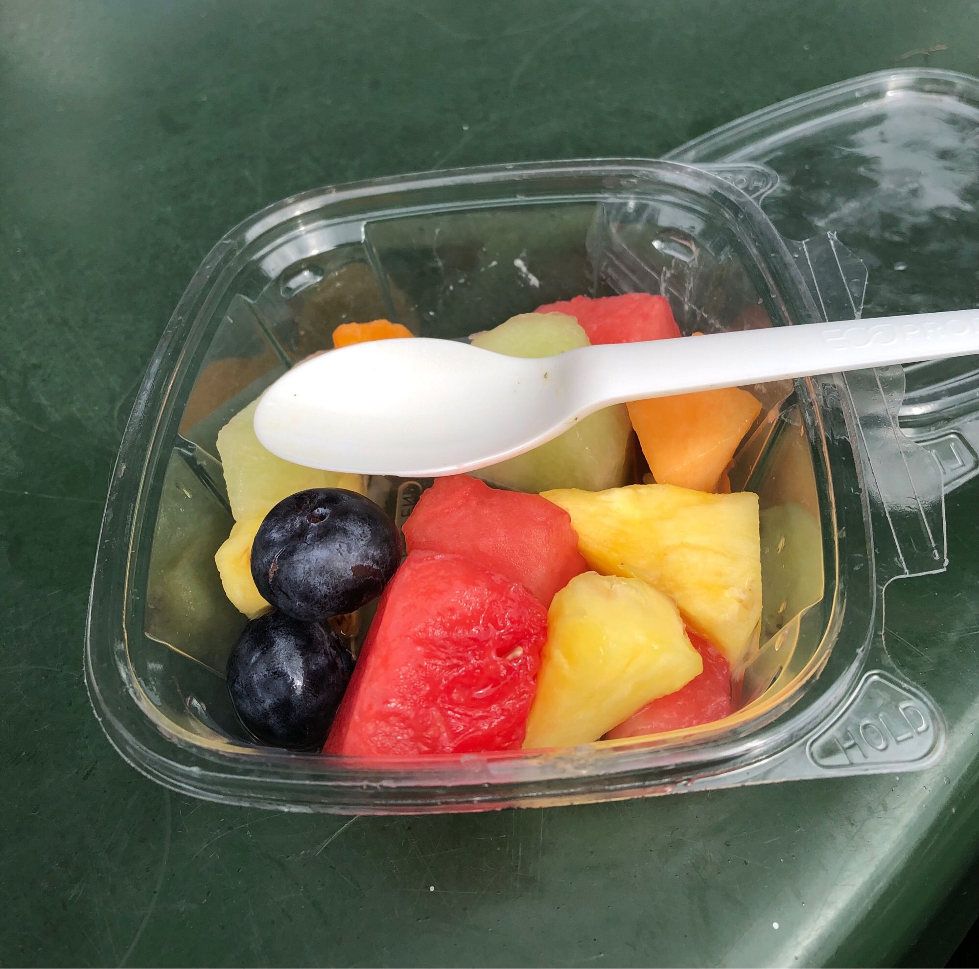 Two huge, dark blueberries in a plastic container among chunks of melon and pineapple. A spoon is laid on top of the salad for scale.