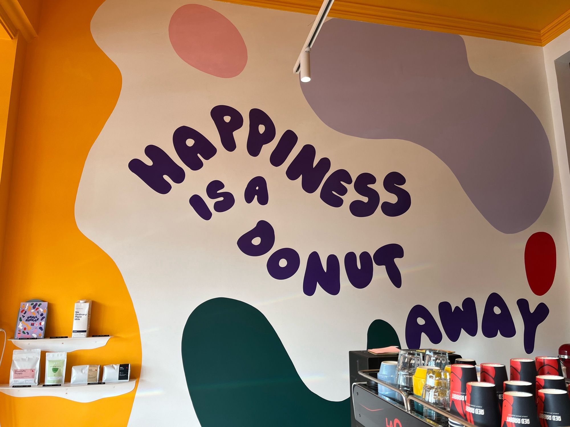 Picture of a vibrant cafe wall with a peak of an espresso machine in the foreground. On the wall is written ‘Happiness is a donut away’.